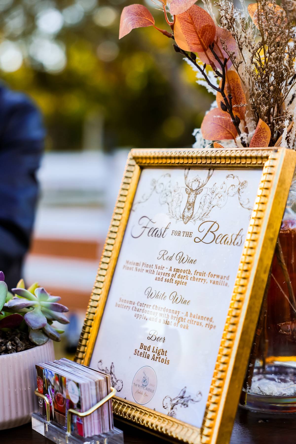 A decorative menu board with gold frame, listing wines and beers, set on a table with succulents in a small pot and a container of sugar packets, with blurred background and leaves in autumn colors.