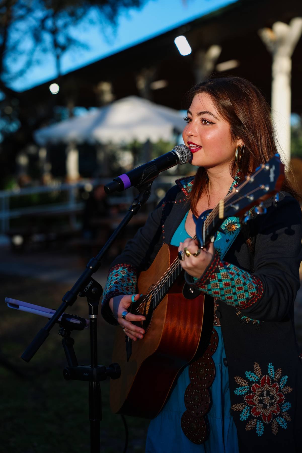 A woman with brown hair playing an acoustic guitar, singing into a microphone, outdoors during evening, with a white umbrella and a dark structure in the background.