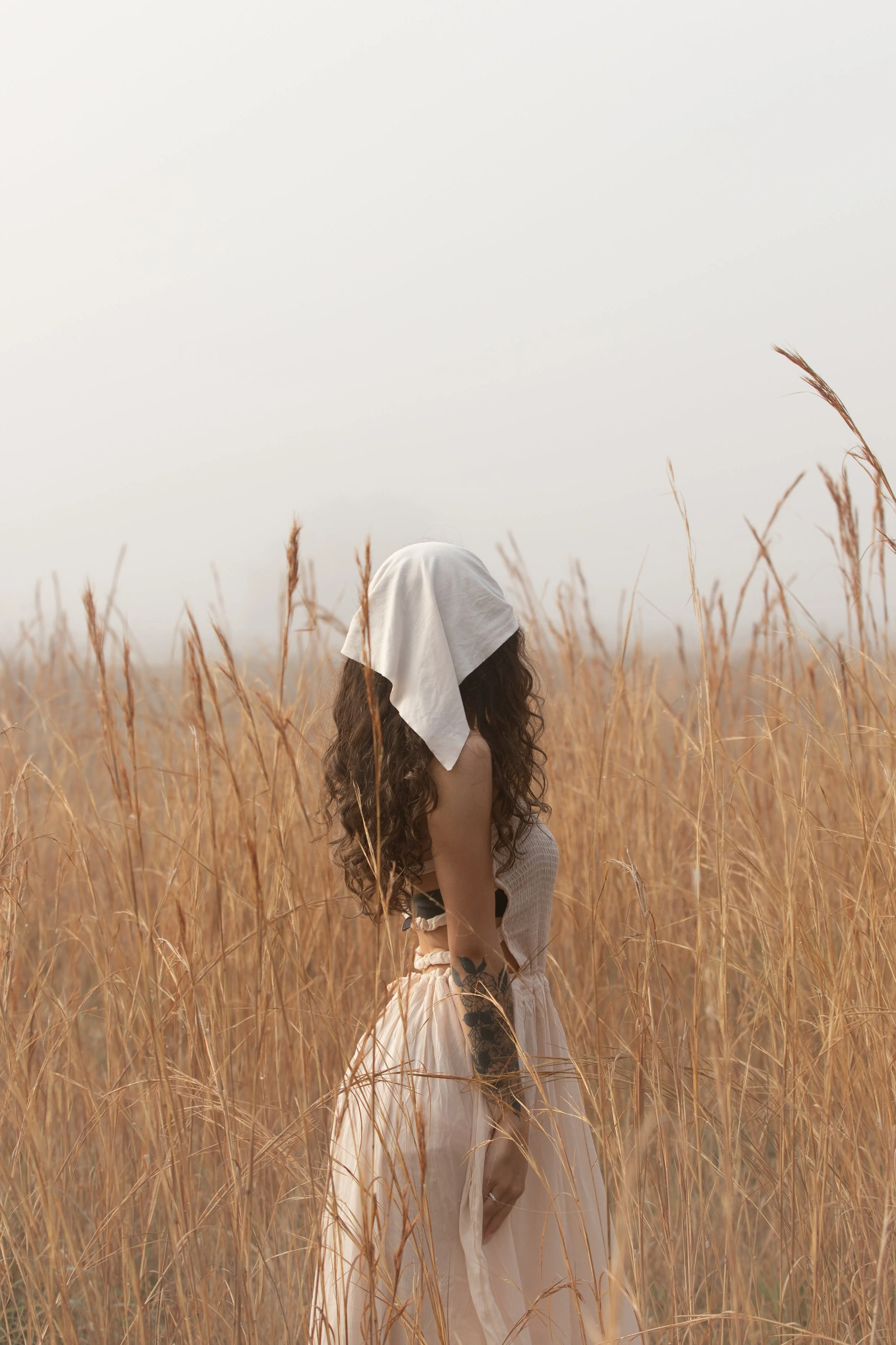 A person with long curly hair wearing a white headscarf sitting in a field of tall, dry grasses.