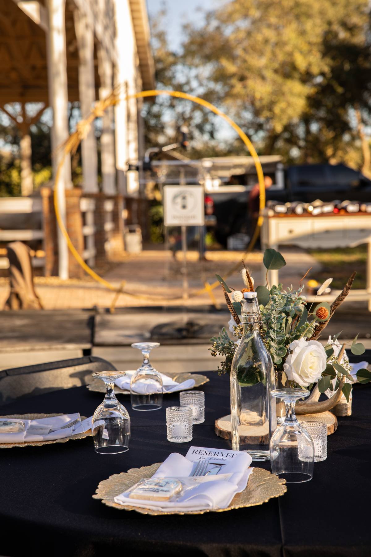 Outdoor dining table decorated with a black tablecloth, white plates, napkins, glassware, and a floral centerpiece with white roses and greenery, set for a special event in a rustic outdoor setting.