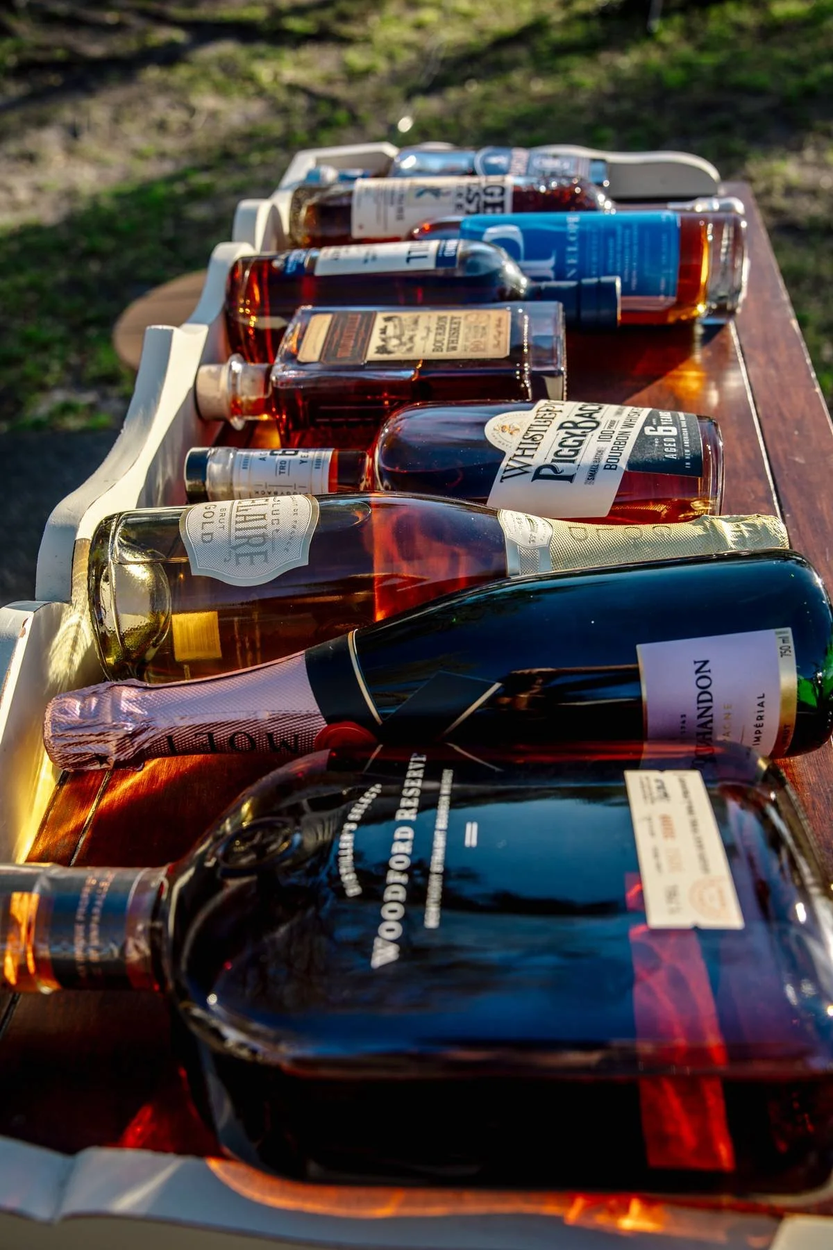A tray of various bottles of alcohol, including whiskey, tequila, and champagne, arranged outdoors on a wooden surface in natural sunlight.