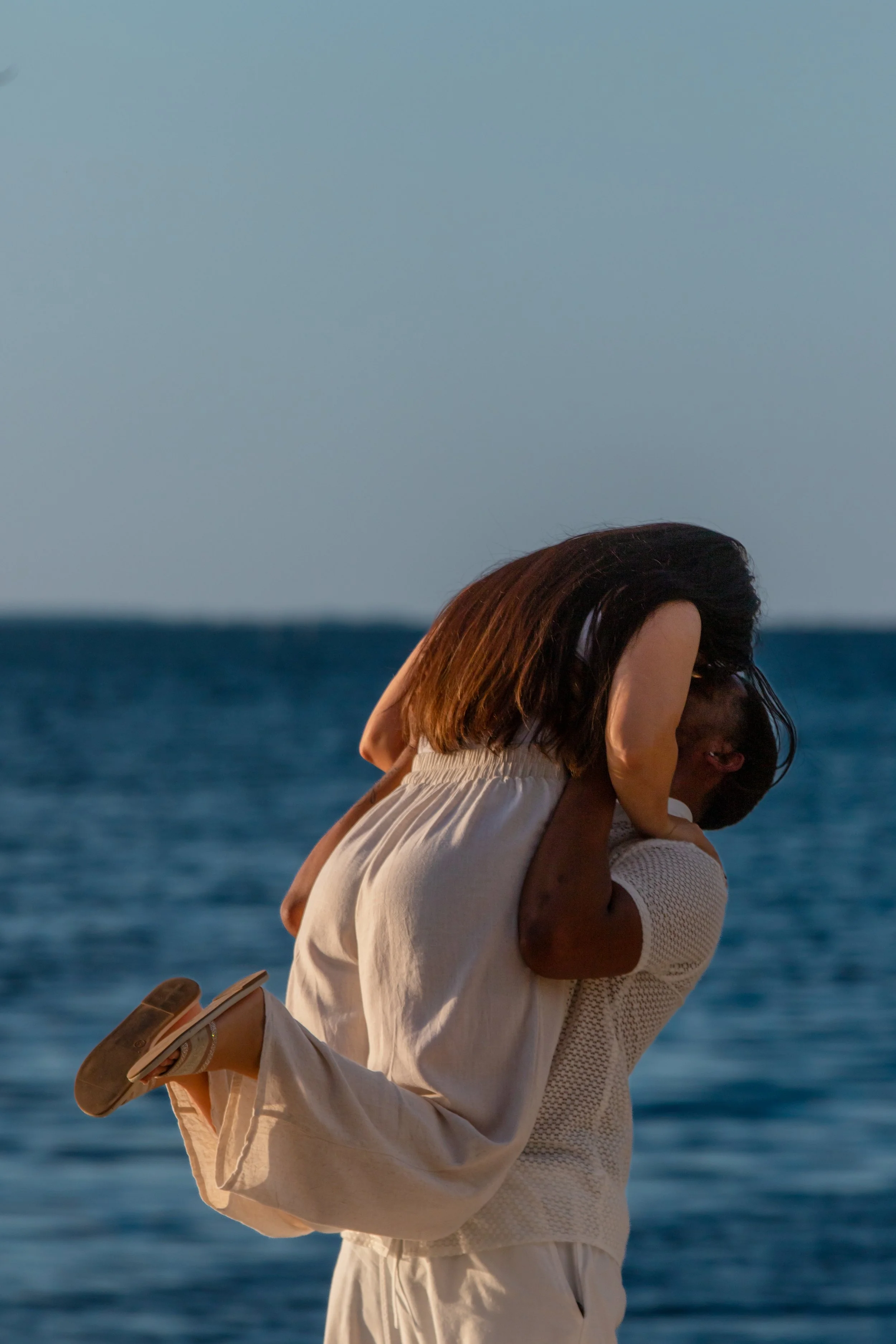 A couple embracing and kissing on the beach near the ocean at sunset, with a woman in a white dress and man wearing a white shirt and beige pants.