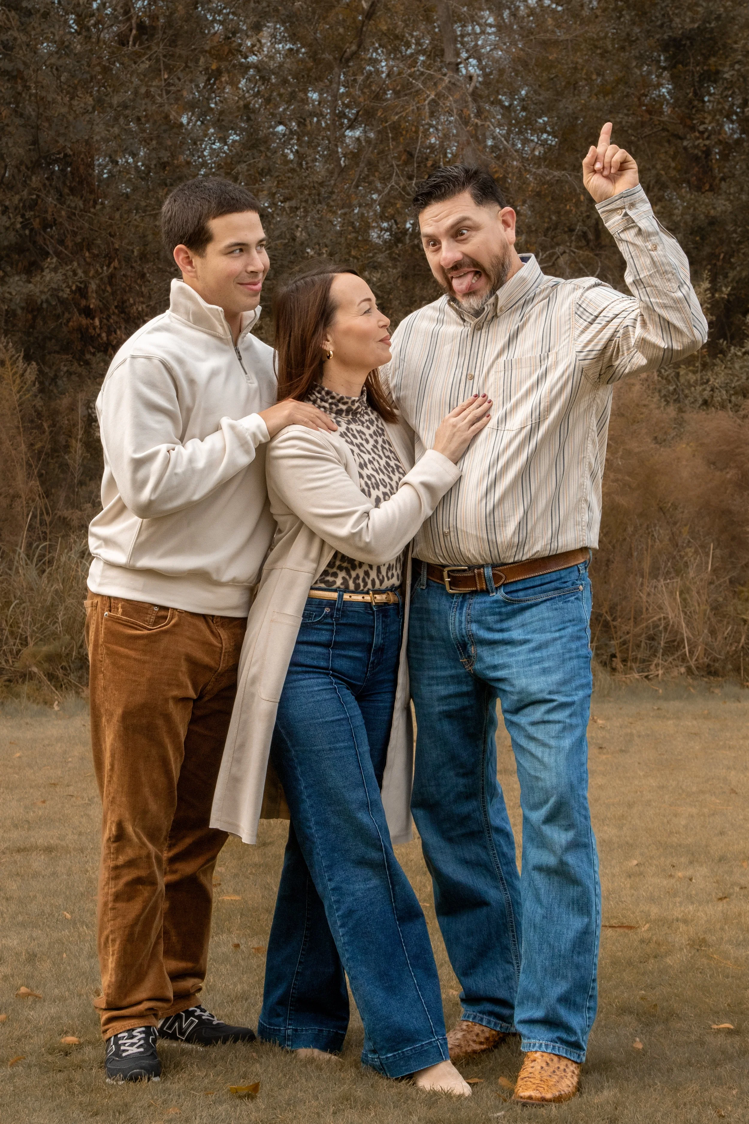 Four people standing outdoors on a grassy field in autumn, with brown trees in the background. They are posing playfully: one man is sticking out his tongue and raising one finger, a woman is smiling and touching his chest, another young man is looki