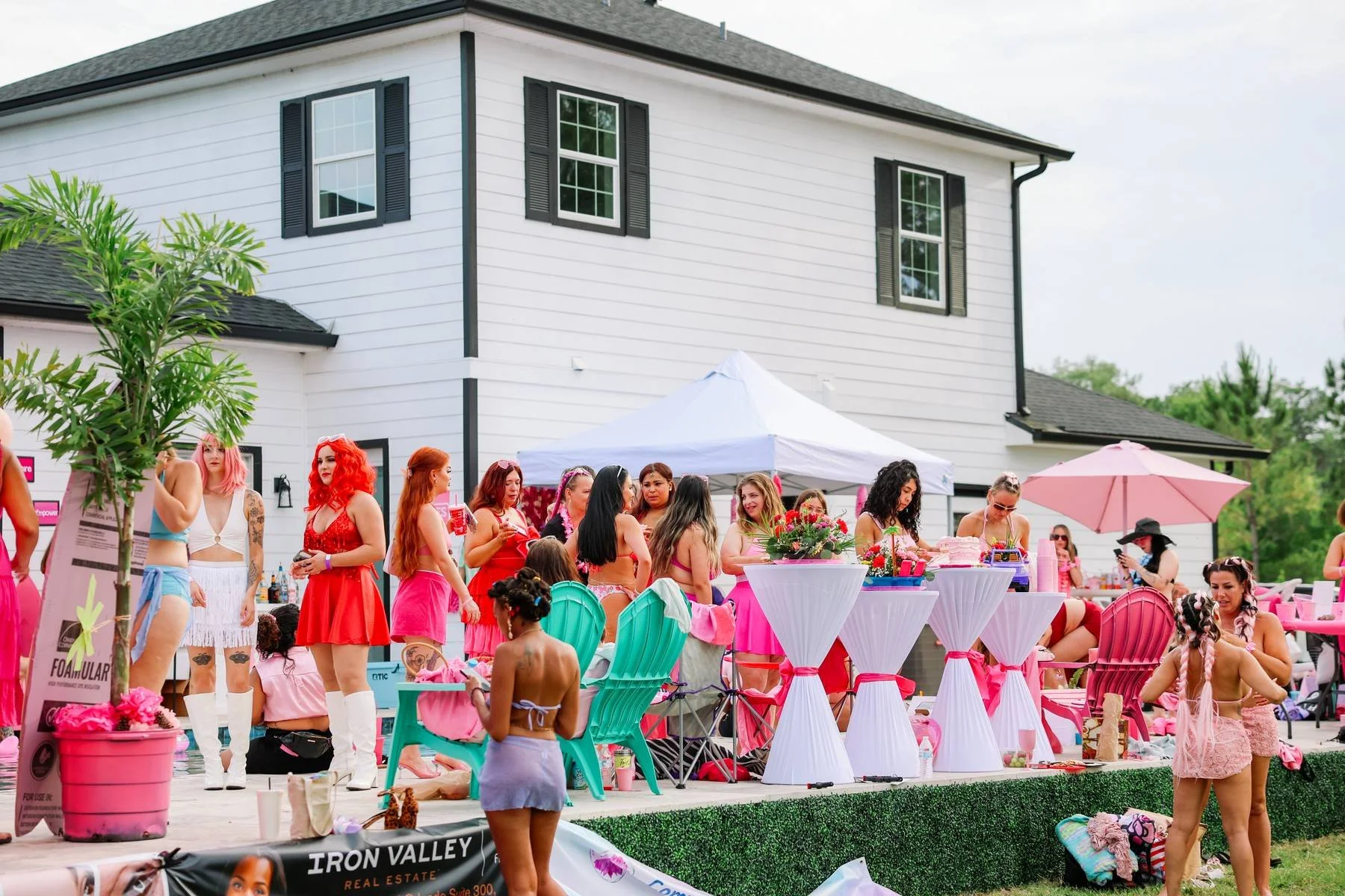 A gathering of women dressed in colorful outfits at an outdoor party, with many seated at tables, and others standing or walking around, set in a residential backyard with a white house in the background.