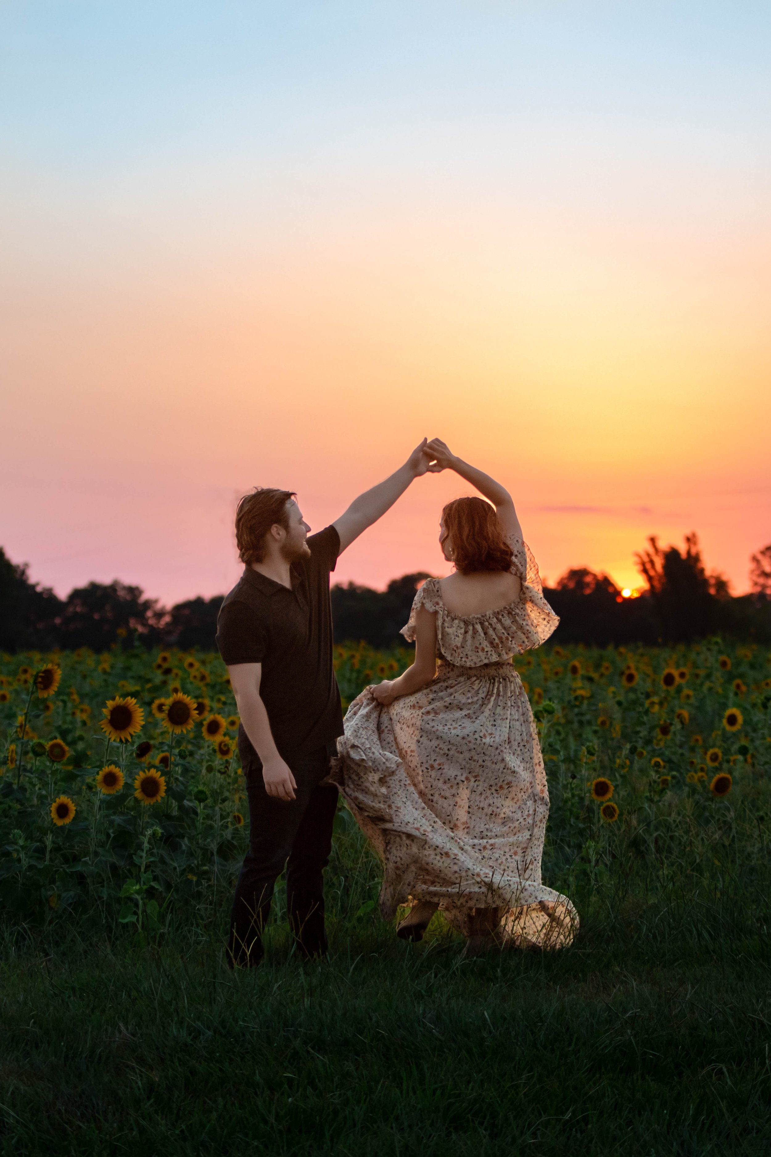 A man and a woman dance in a field of sunflowers during sunset, with the man lifting the woman by the hand in a romantic scene.