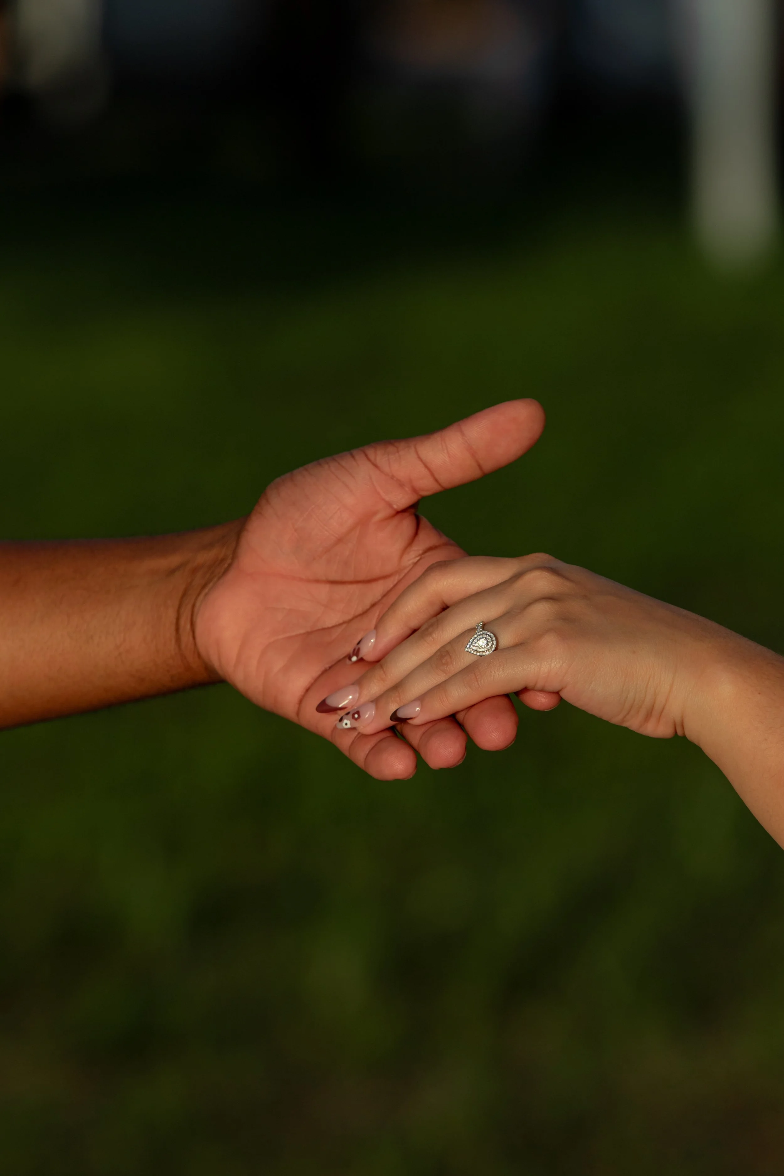 Close-up of a bride and groom holding hands, with the bride wearing a ring, against a blurred outdoor background.