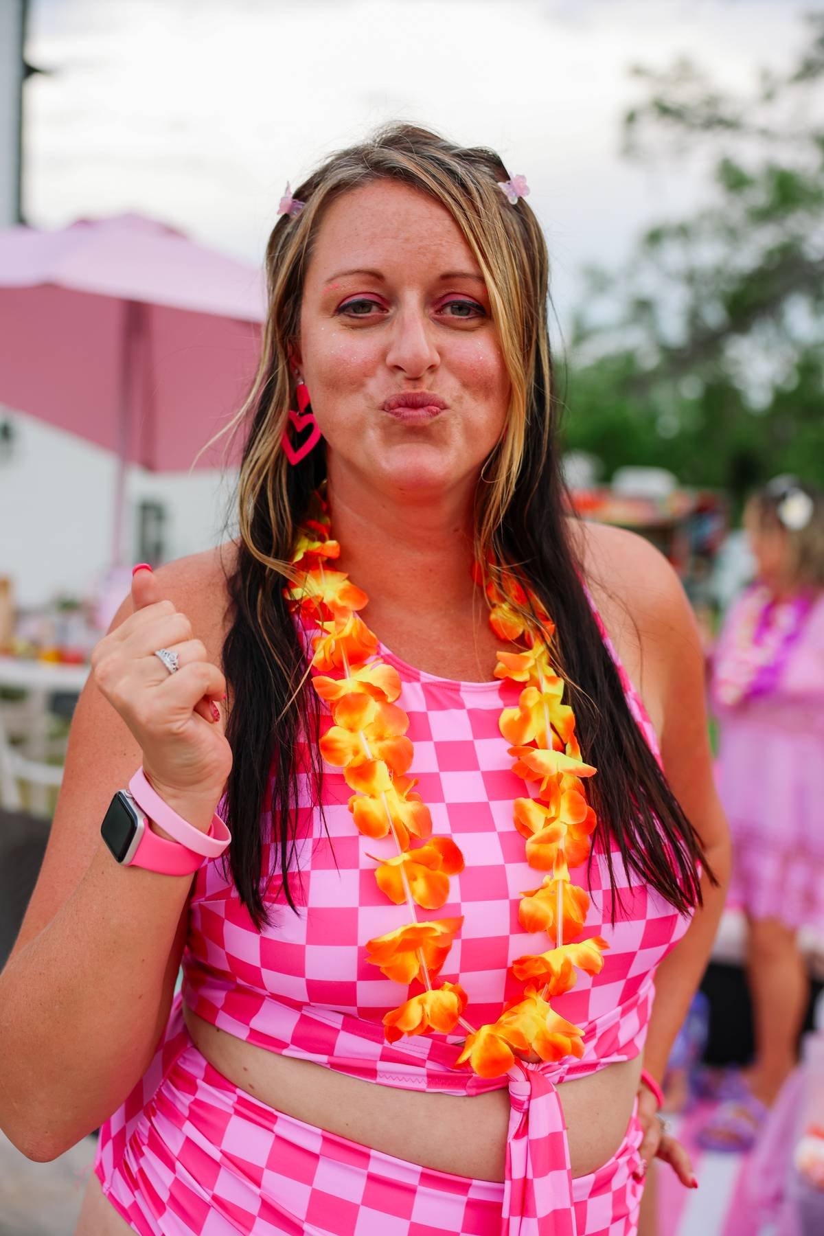 A woman wearing a pink checkered swimsuit, a flower lei, and pink heart earrings, celebrating at a tropical-themed event outdoors.