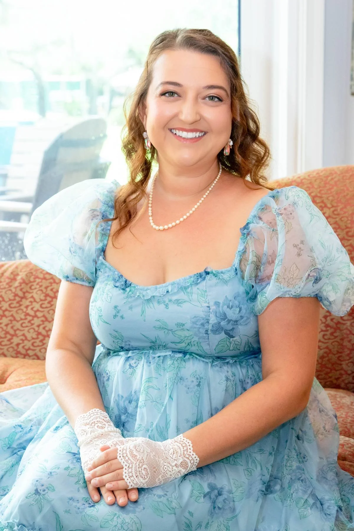 A smiling woman wearing a light blue floral dress, pearl necklace, and lace gloves, sitting on a sofa in a brightly lit room with large windows.