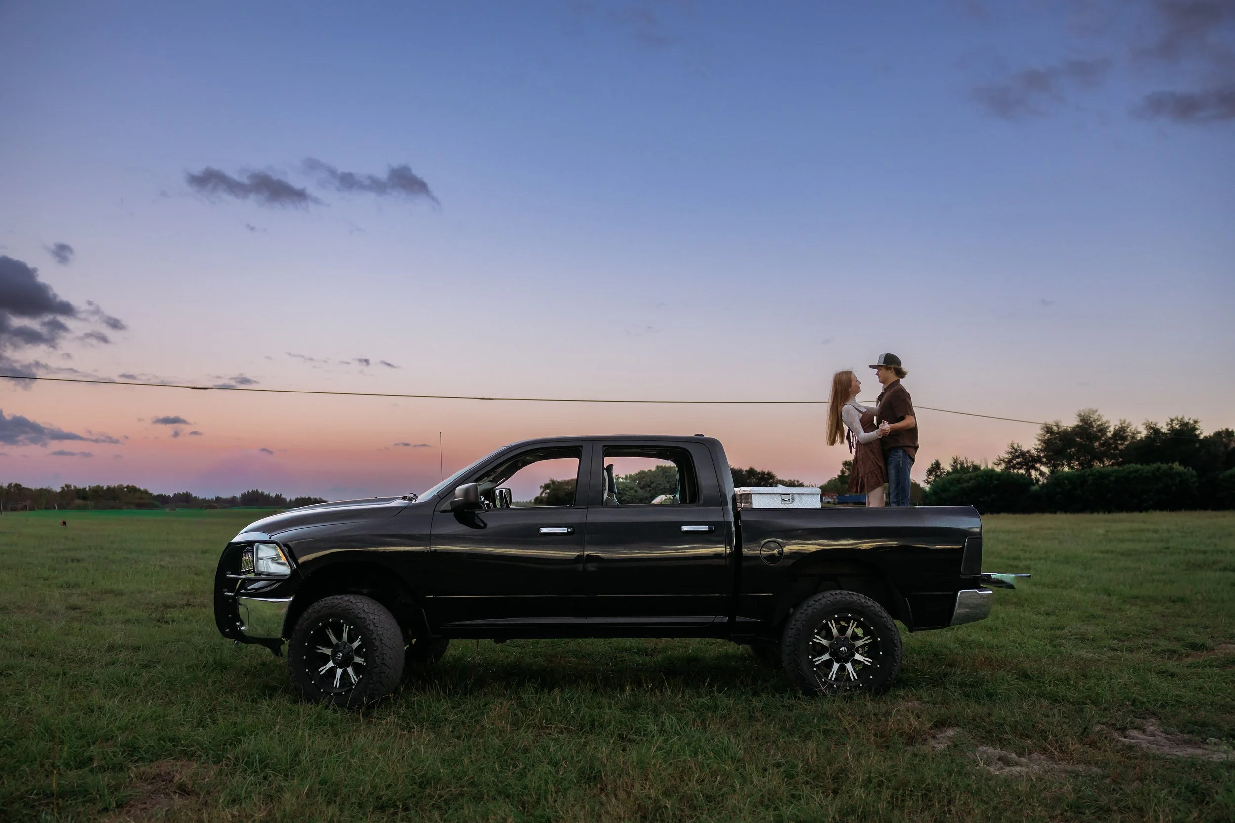 A black pickup truck parked on grass with a couple standing behind it, holding hands and smiling at each other during sunset.