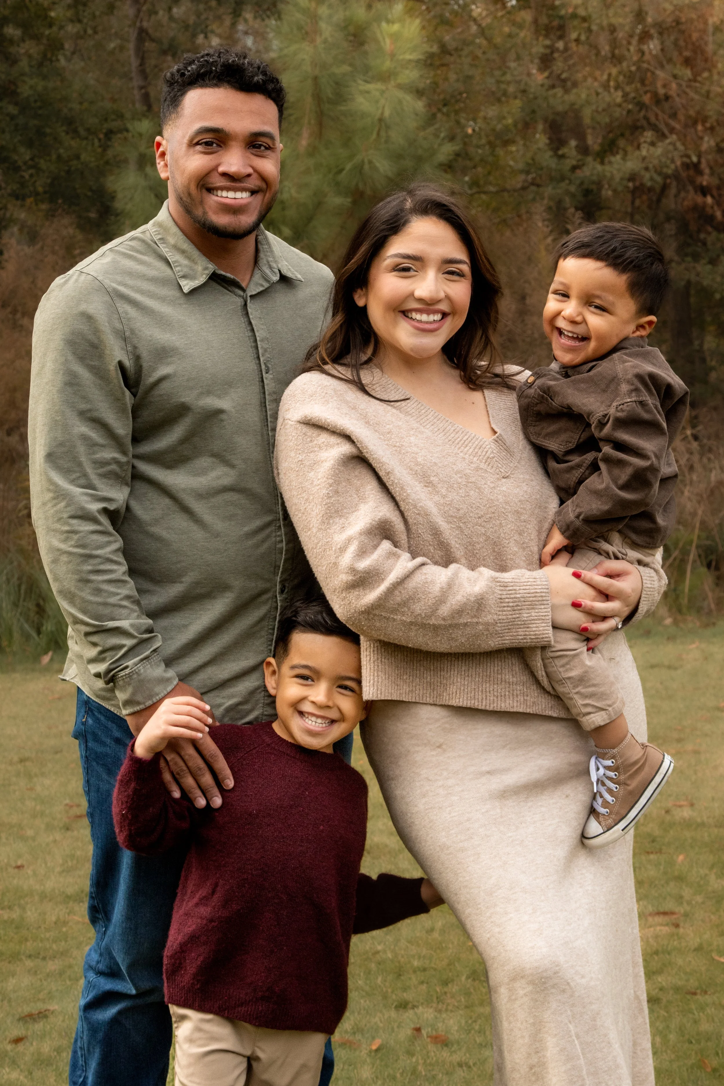 A family of five, including two children, smiling outside during fall with trees and grass in the background.