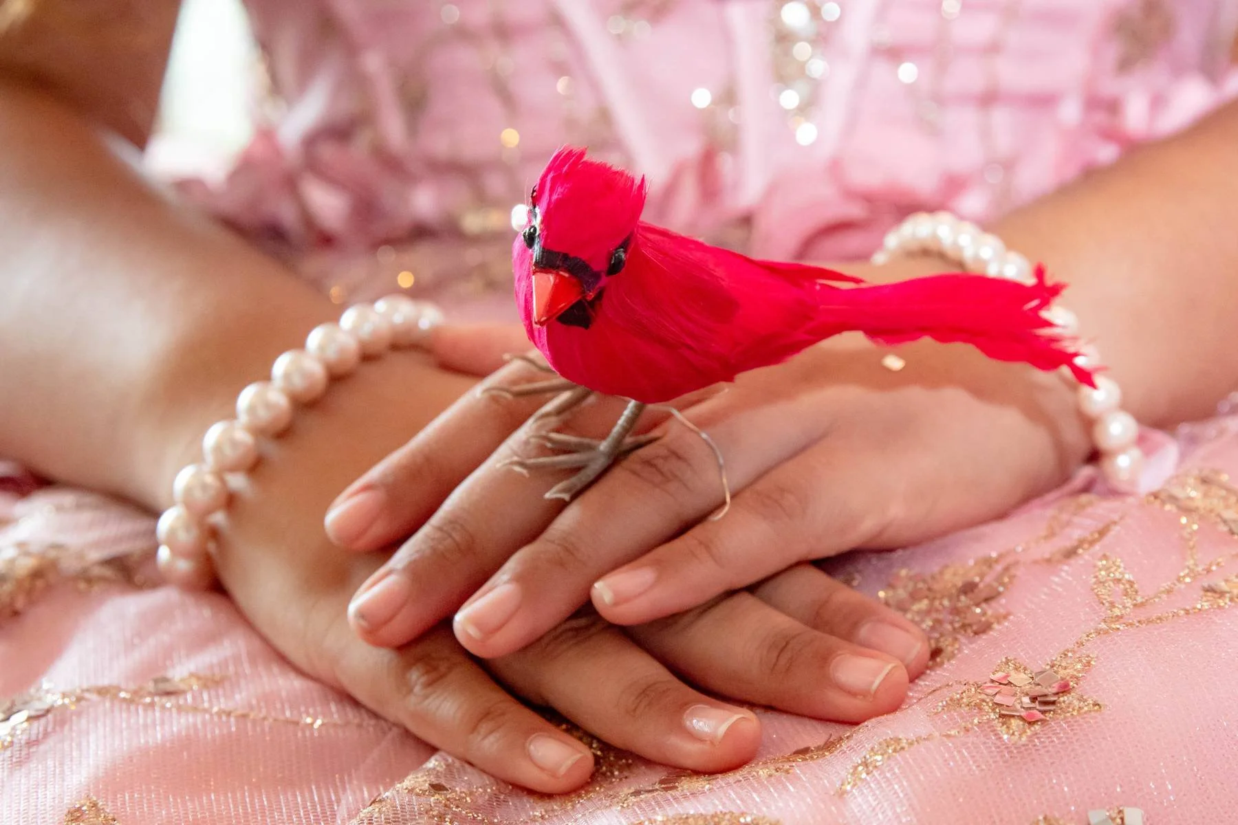 A pink artificial bird perched on a person's finger, with jewelry like pearl bracelets and a pink, glittery, textured dress in the background.