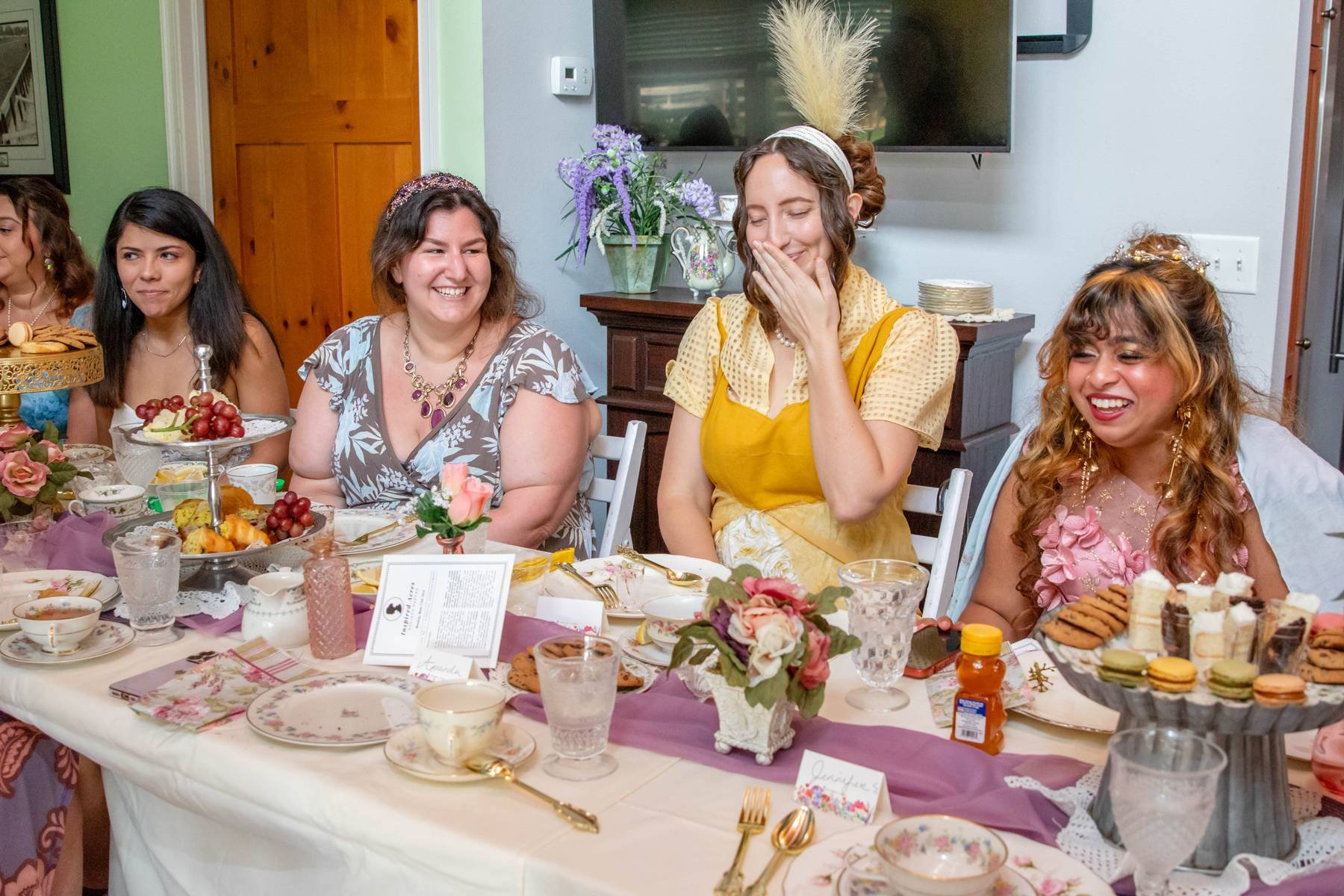 A group of women at a celebration, sitting around a table with food and decorations, some wearing tiaras and hats, smiling and laughing.
