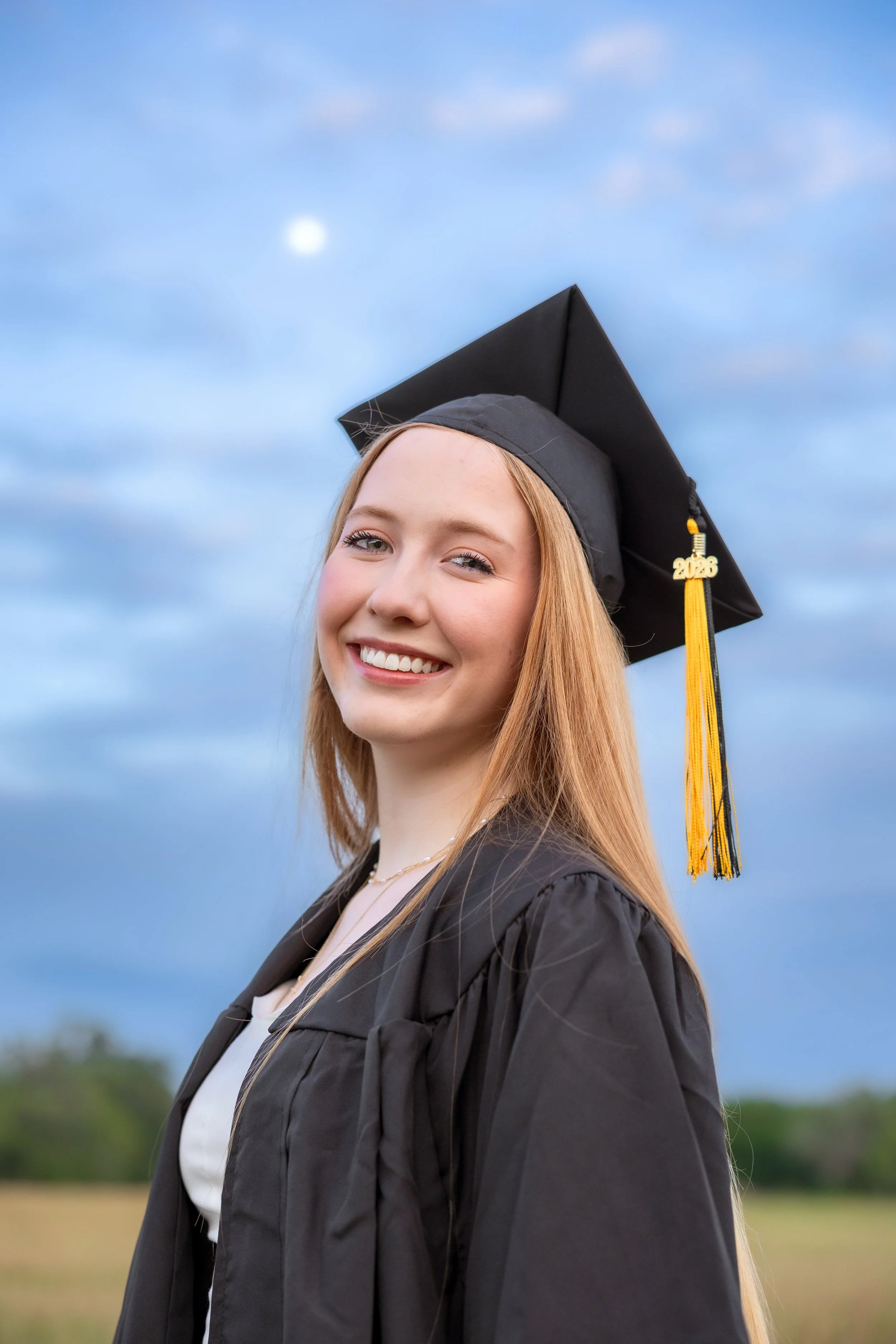 A young woman with long blonde hair smiling outdoors during graduation, wearing a black cap and gown with a yellow tassel marked '2025', against a blue and partly cloudy sky.