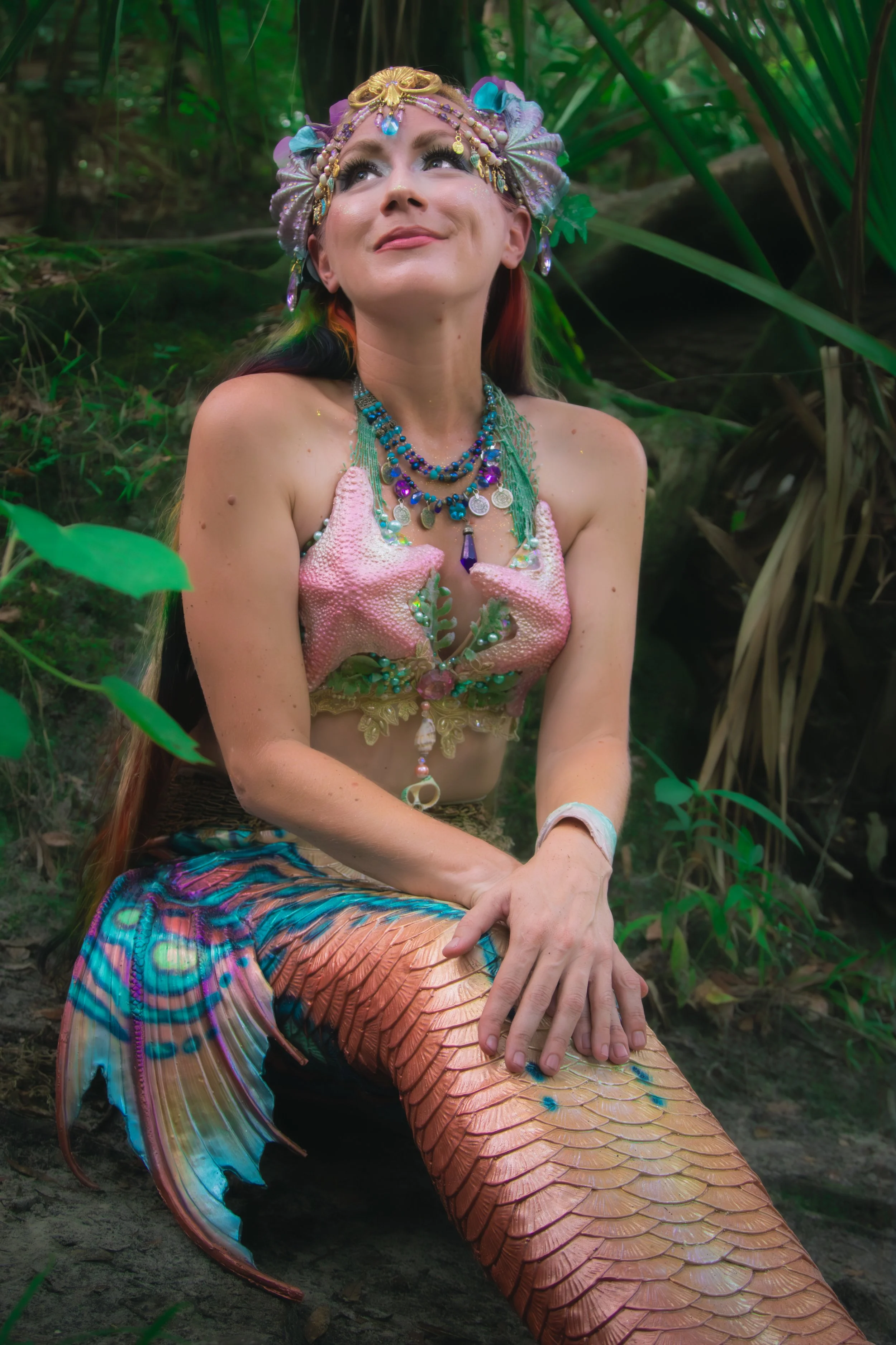 A woman dressed as a mermaid, with a seashell top, colorful jewelry, and a mermaid tail, sitting outdoors in a lush green jungle-like environment.