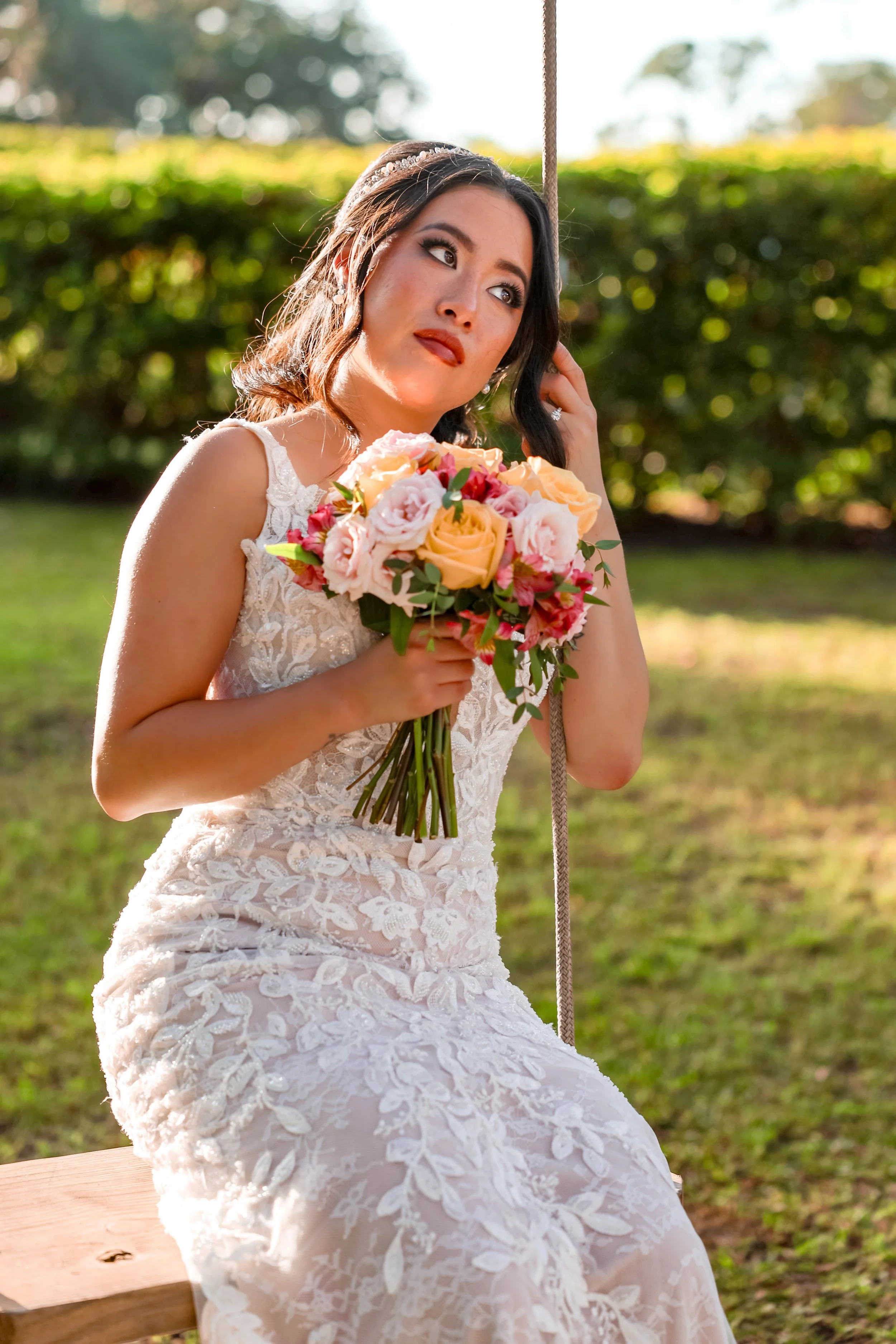 A woman in a lace wedding dress sitting on a wooden bench outdoors, holding a bouquet of pink and peach roses, with greenery and trees in the background.