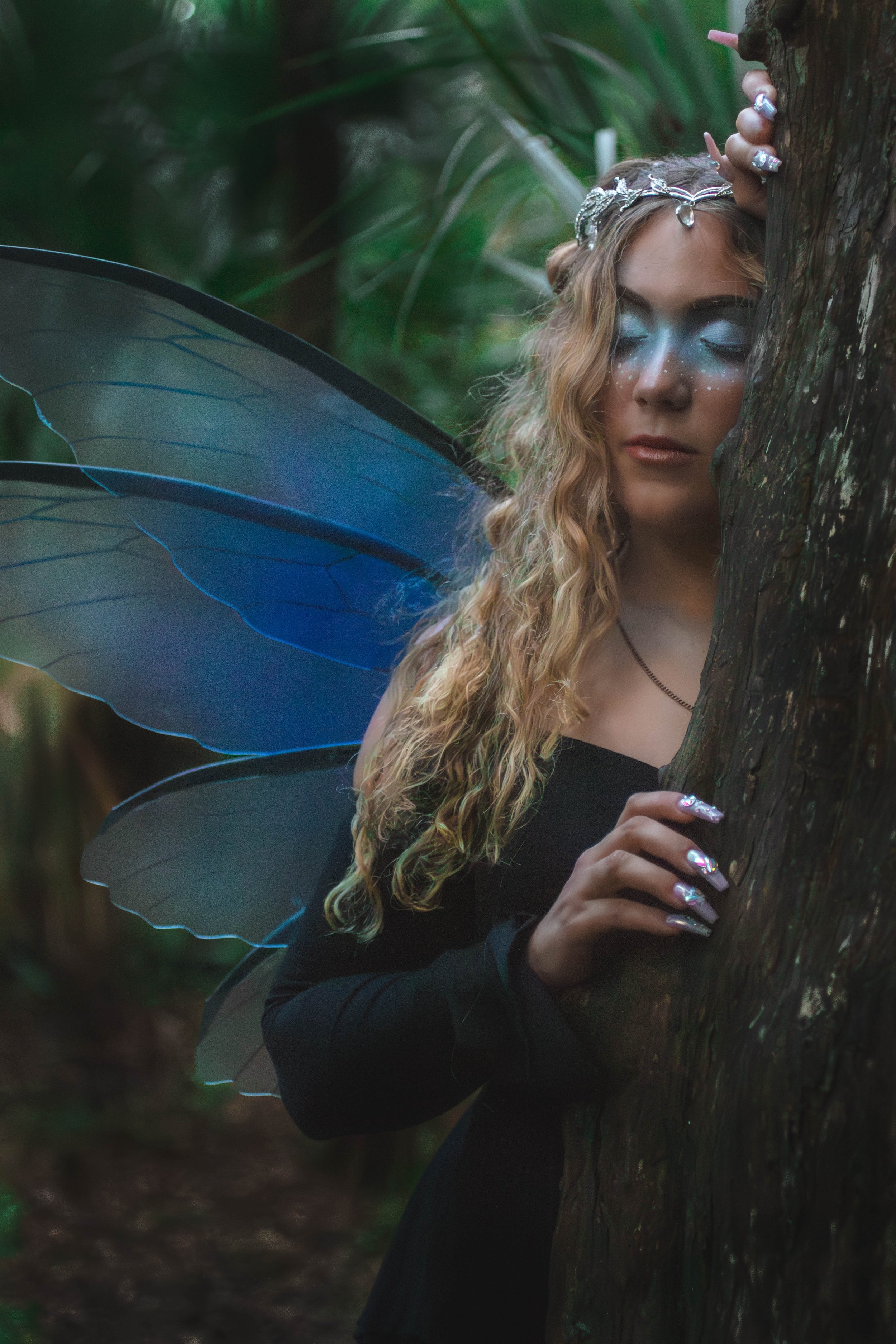 A woman dressed as a fairy or elf with blue butterfly wings, silver jewelry, and metallic nail polish, standing behind a tree in a forest with her eyes closed and a peaceful expression.
