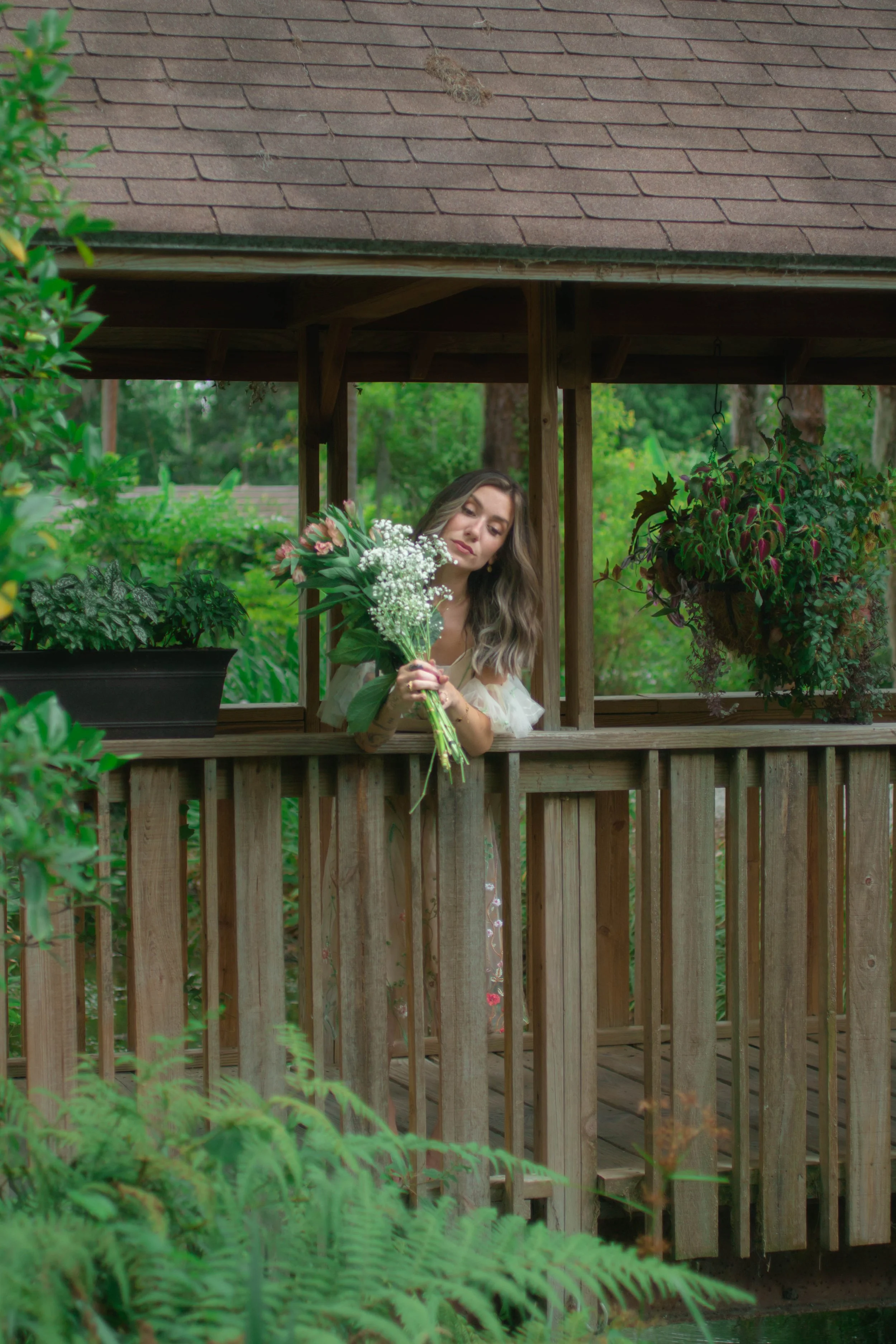 A woman holding a flower bouquet on a wooden porch surrounded by green plants and hanging flower baskets.