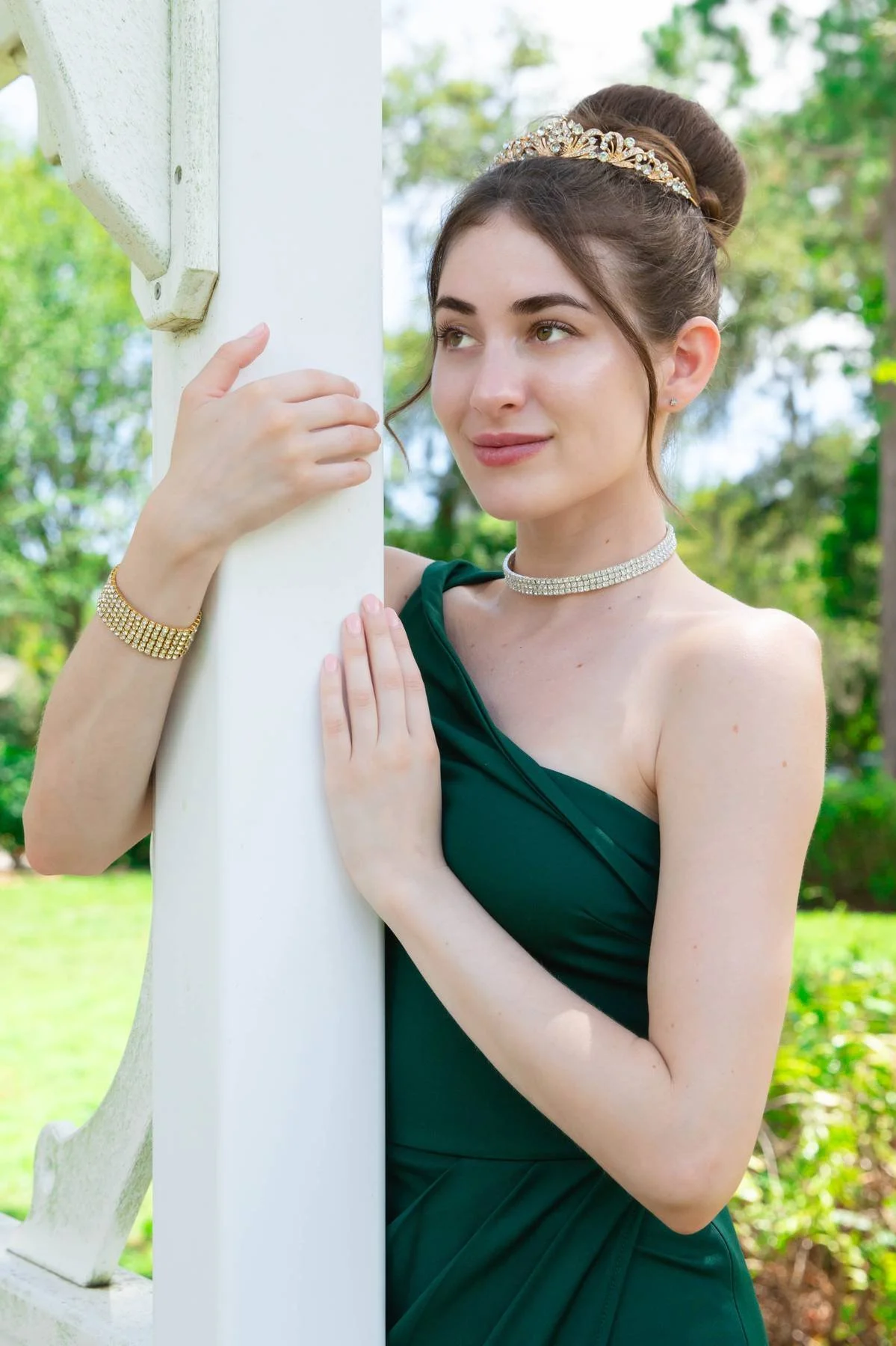 Young woman in a dark green dress and jewelry, embracing a white wooden post outdoors with trees and greenery in the background.