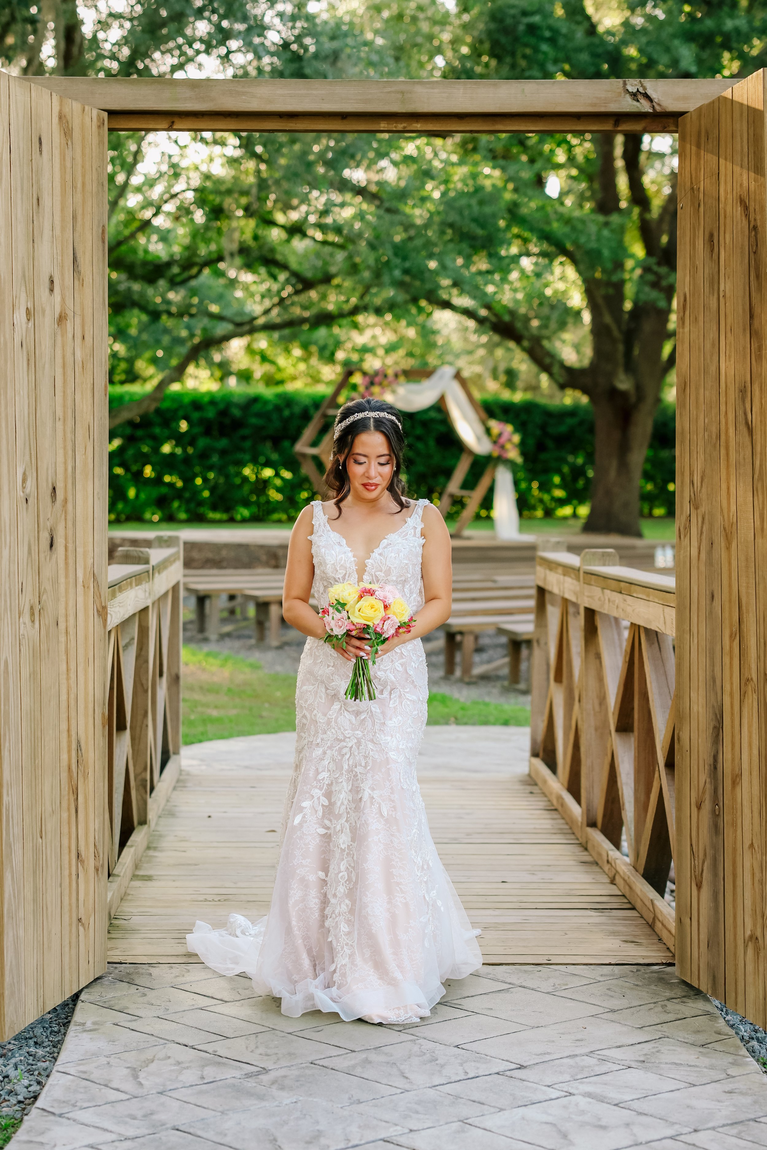 A bride in a white lace wedding dress holding a bouquet of yellow, pink, and white flowers standing on a wooden bridge with a natural outdoor setting, trees, and green foliage in the background.