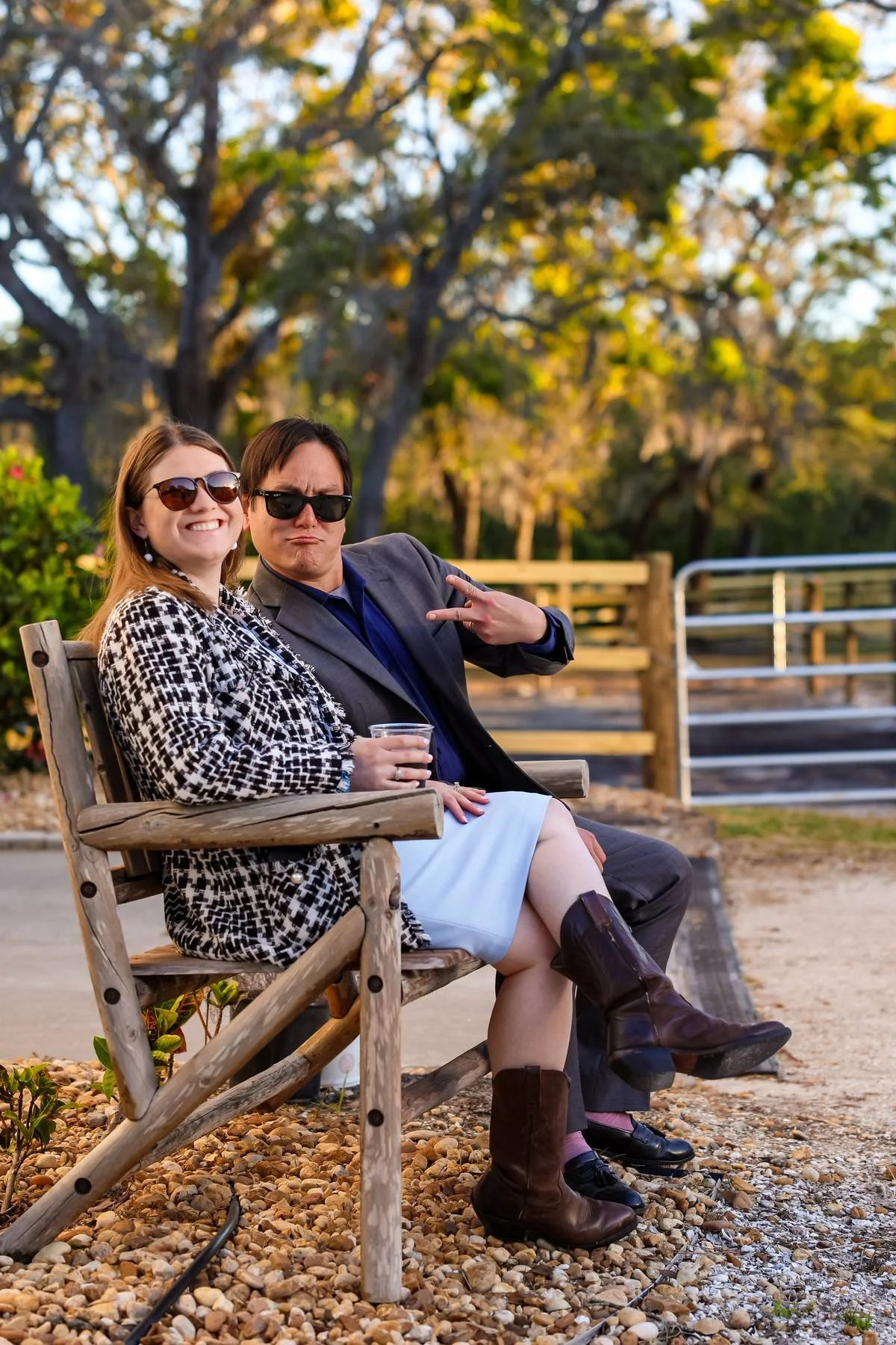 A man and woman sitting on a park bench, smiling and making playful gestures, with trees in the background during daytime.