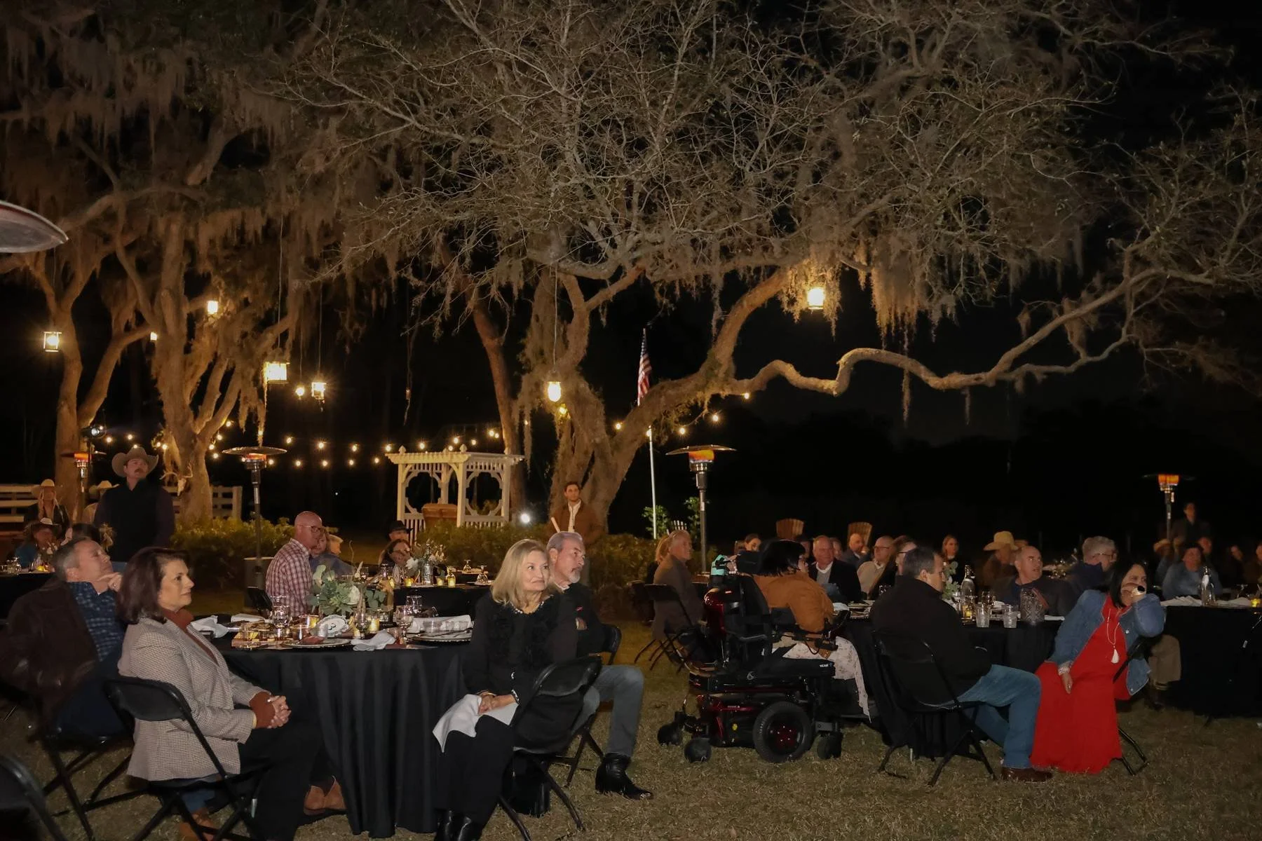 People attending an outdoor evening event seated at round tables with black tablecloths, surrounded by large trees decorated with hanging lanterns, with some individuals in wheelchairs and others standing or sitting, under string lights and lanterns 