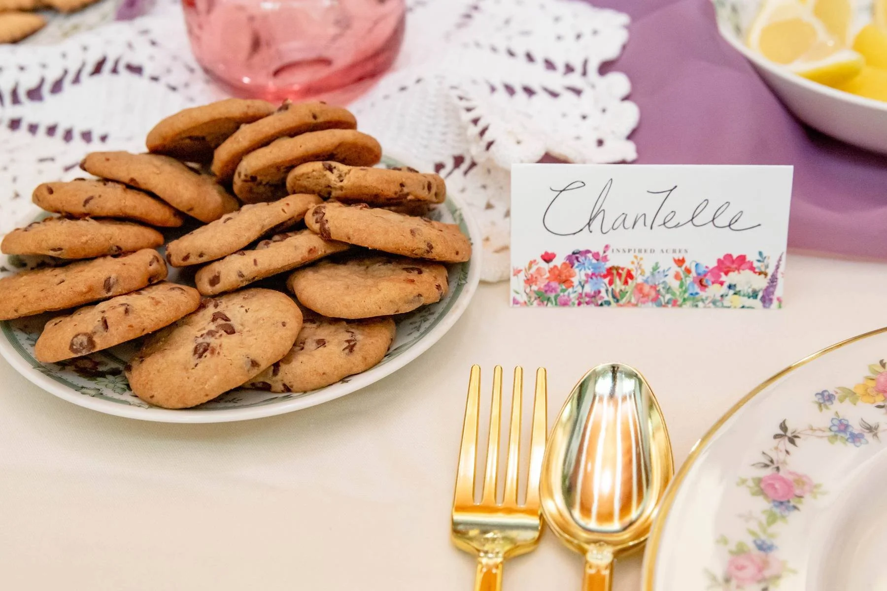 Plate of chocolate chip cookies on a lace doily with a name card that says 'Chantelle' and a floral design, gold utensils, and a floral plate partially visible.