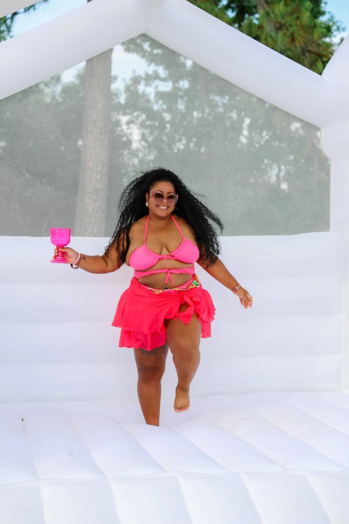 A woman in a pink bikini top and red skirt holding a pink glass and smiling, standing on an inflatable white pool float with a mesh hut background and trees.