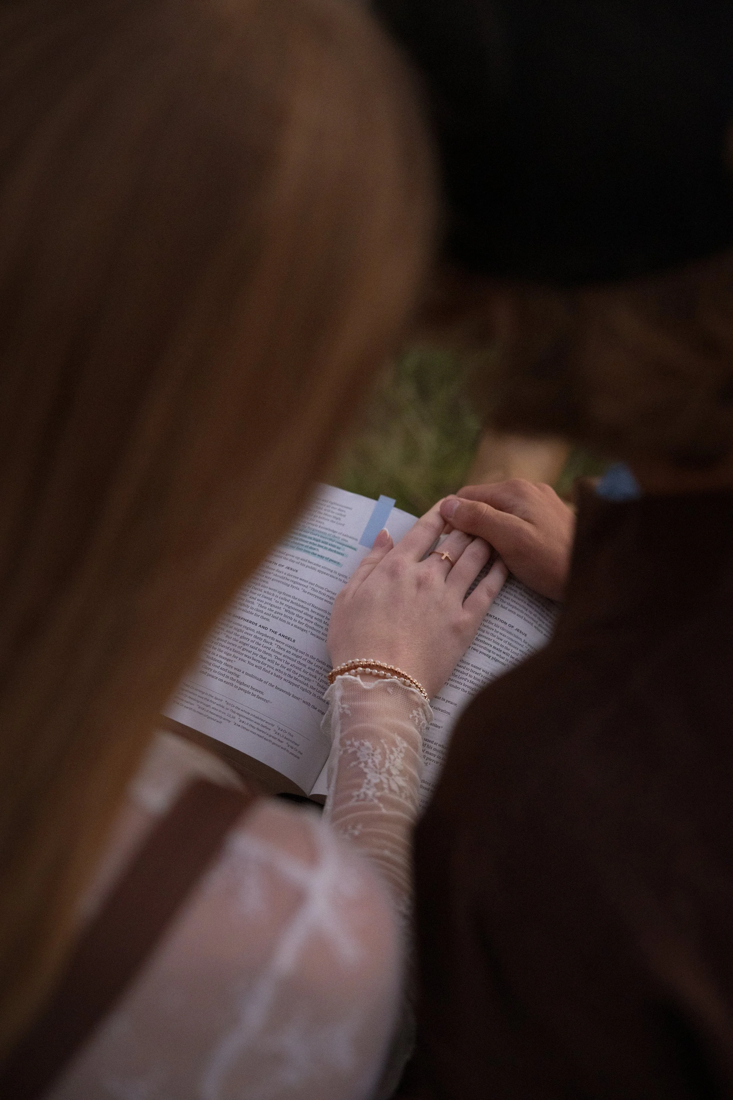 A person in a lace dress is reading a book titled 'Acts and the Angels' outdoors, with another person's hands resting on the page.
