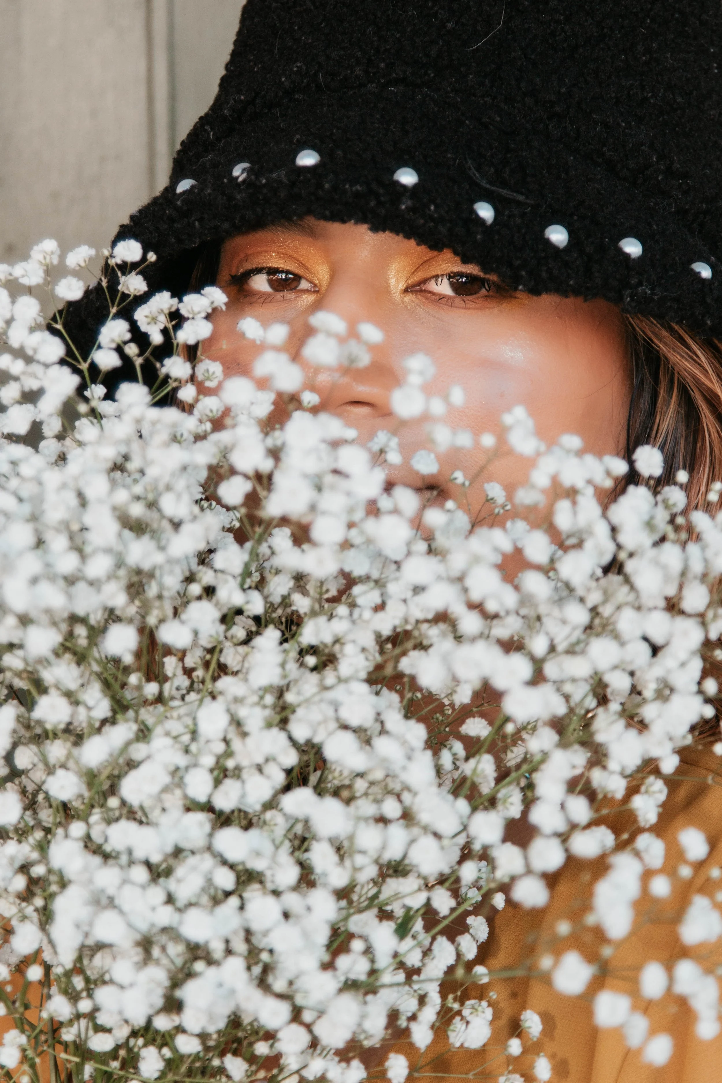 A woman wearing a black beanie hat decorated with pearls, with her face partially obscured by white baby's breath flowers.
