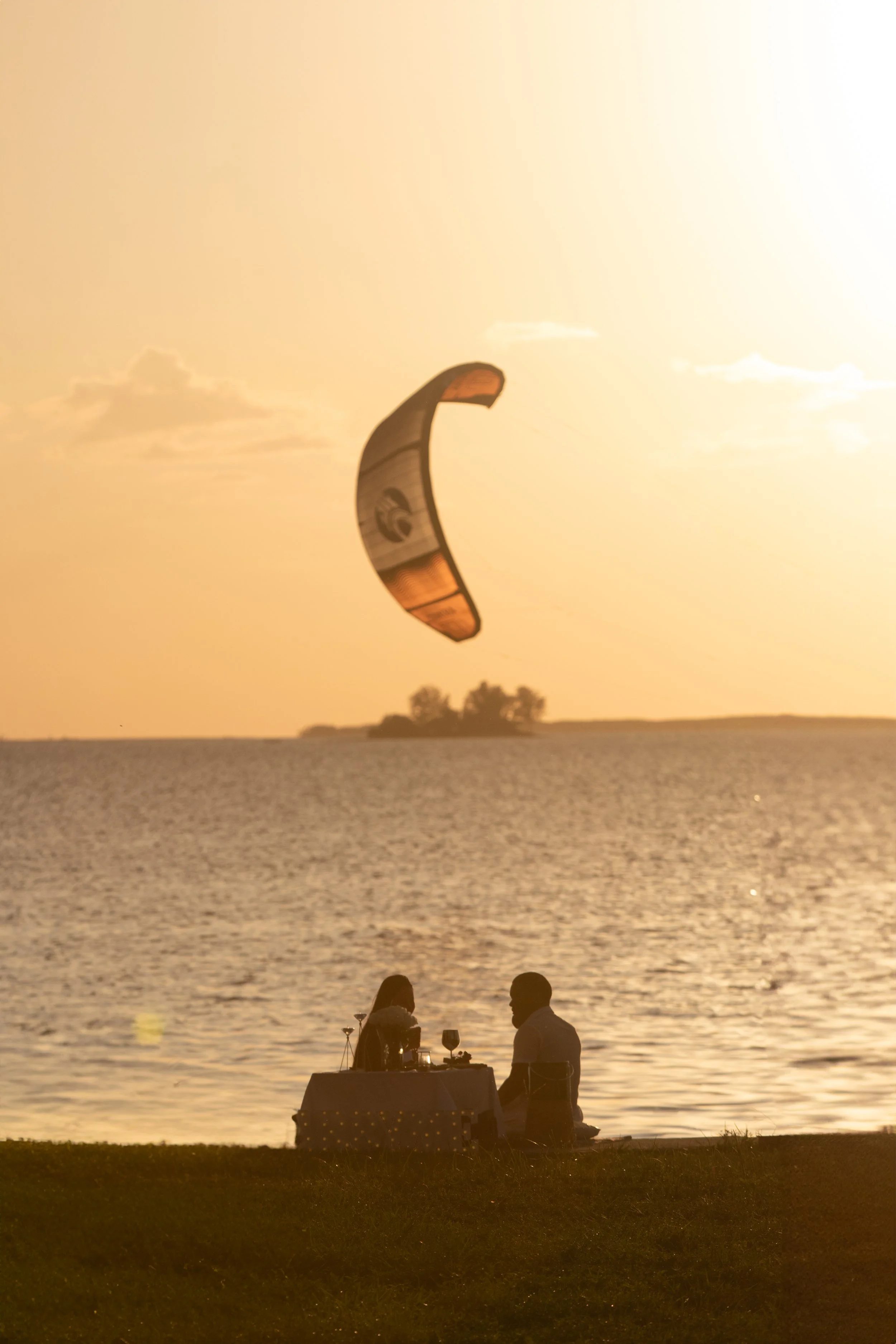 A sunset by a lake with a kite surfer soaring in the sky, and two people sitting at a table near the shore.