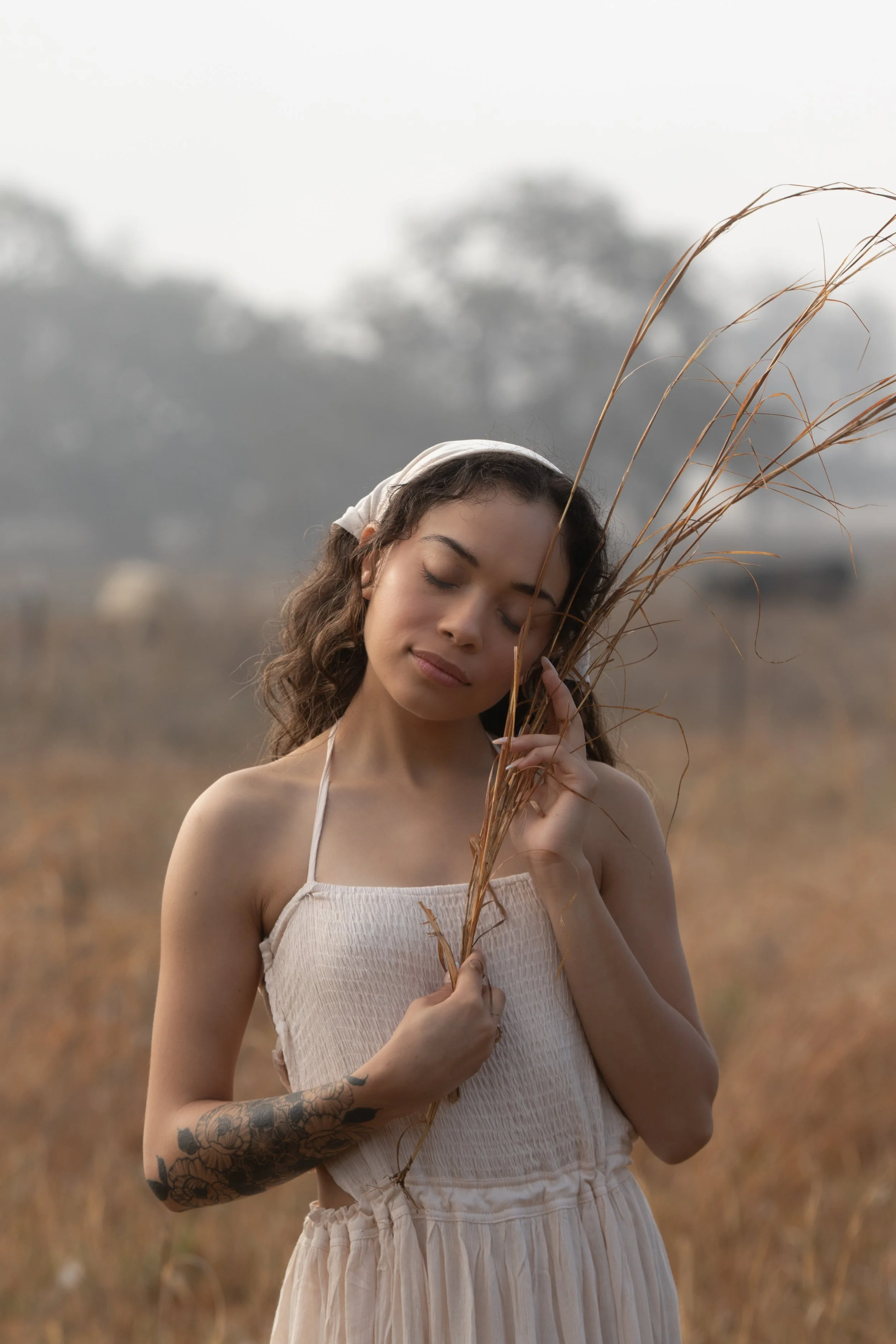 A young woman with brown curly hair and tattoos on her left arm, dressed in a beige dress and a headscarf, standing in a field of tall, dry grass with her eyes closed, holding a bunch of dried grass near her face, with a blurred background of trees a