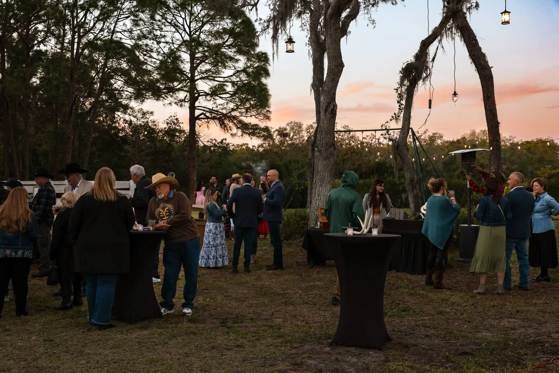 Outdoor social gathering during sunset with guests mingling under trees decorated with hanging lanterns, some wearing hats and others in formal attire, with tables set up for drinks and food.