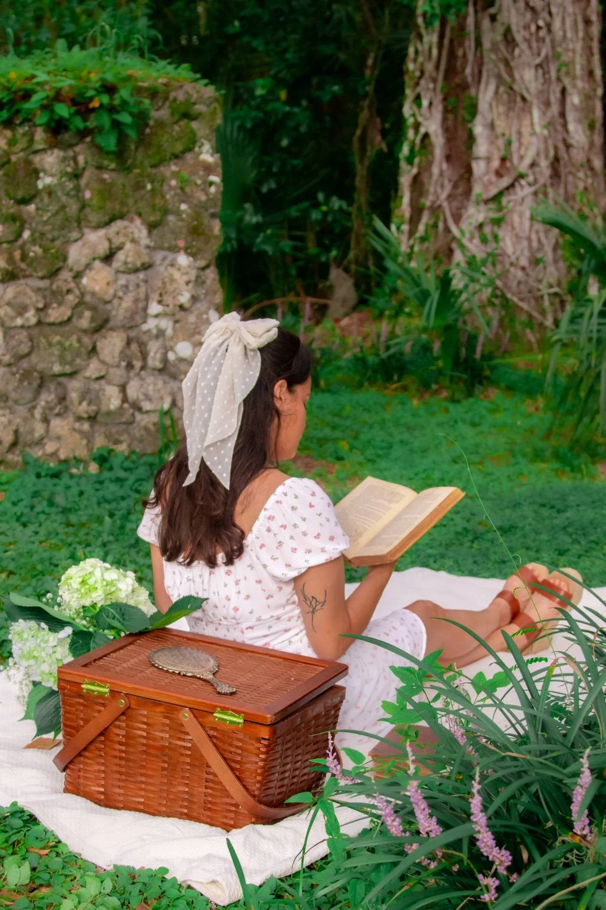 A woman sitting outdoors on a blanket in a garden, reading a book, with a picnic basket and a flower bouquet nearby.
