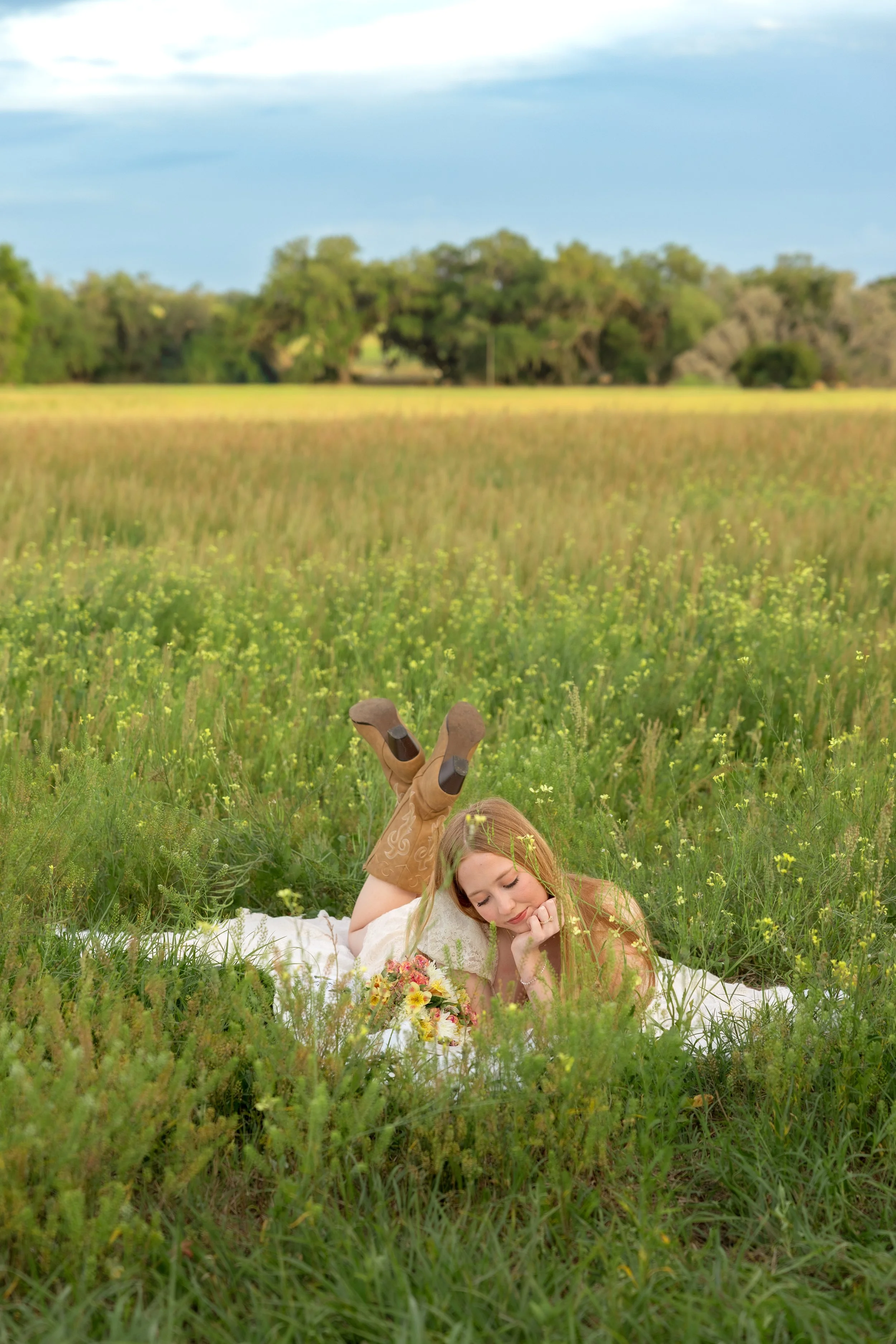 A young woman with red hair lying on her stomach in a grassy field, resting her head on her hand, with a bouquet of flowers nearby, under a partly cloudy sky with trees in the background.