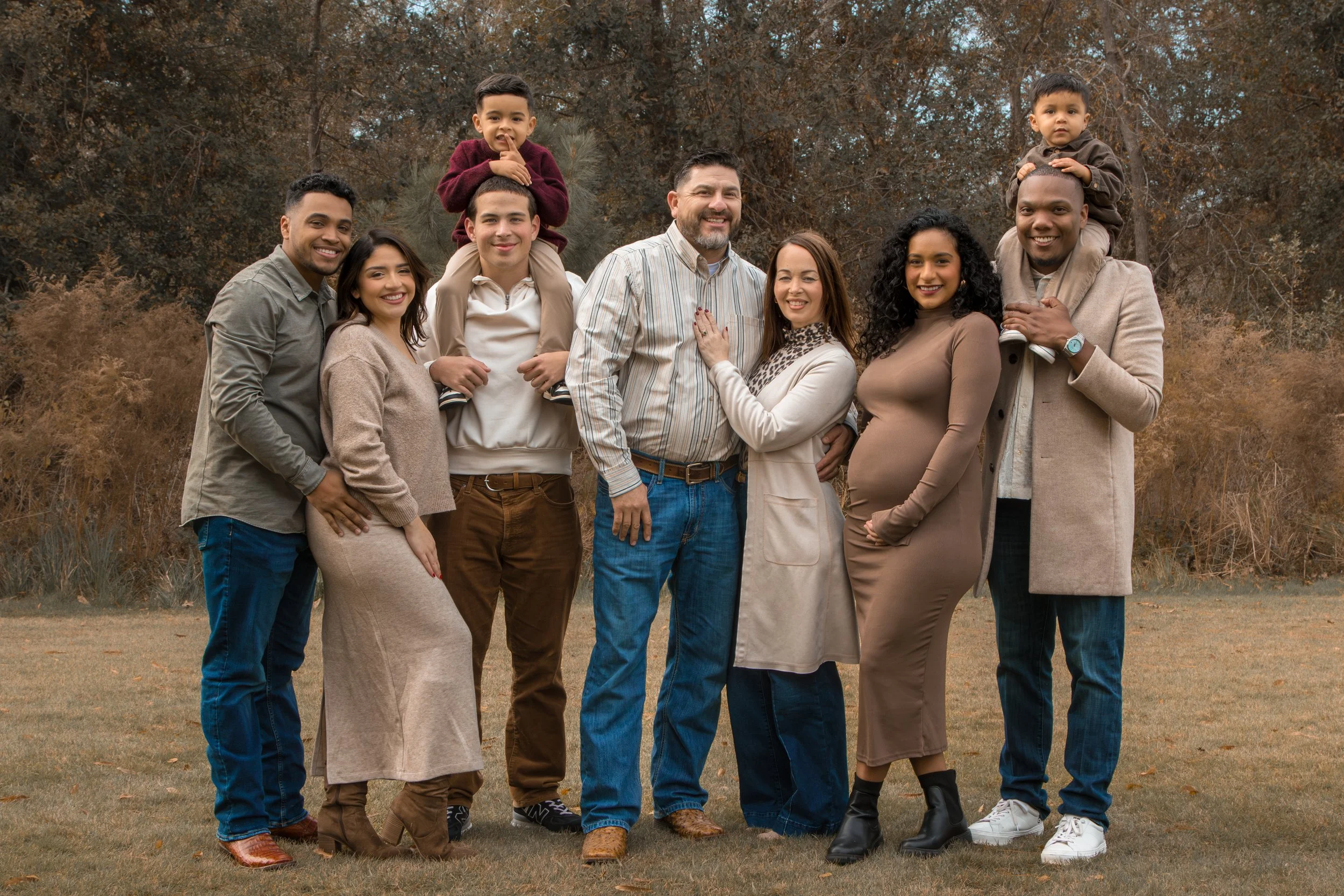 A diverse family of ten people, including children, teenagers, and adults, standing outdoors on a grassy area with trees in the background. They are smiling and posing together for a group photo in an autumn setting.