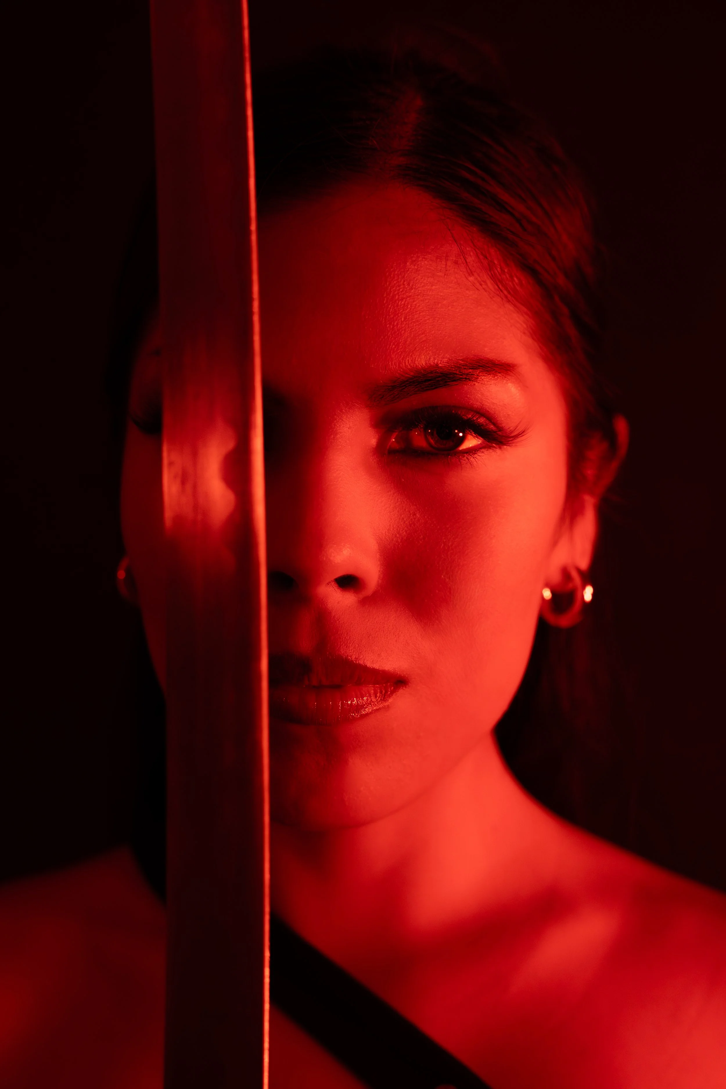 Close-up portrait of a woman with short hair and earrings, divided by a vertical red line, with half of her face illuminated and the other half dark. The lighting creates a mysterious and dramatic effect.