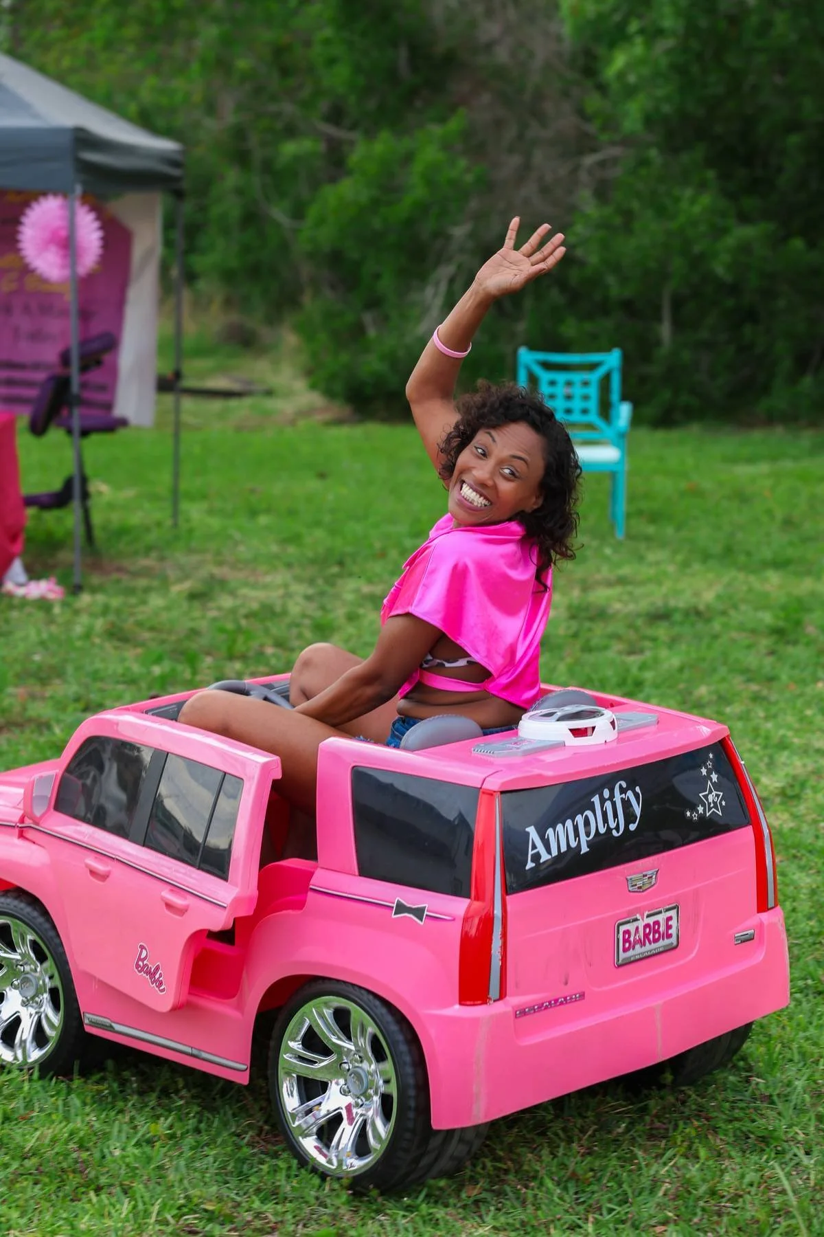 A woman with curly hair, wearing a pink top, sitting inside a pink toy Cadillac, smiling and waving.