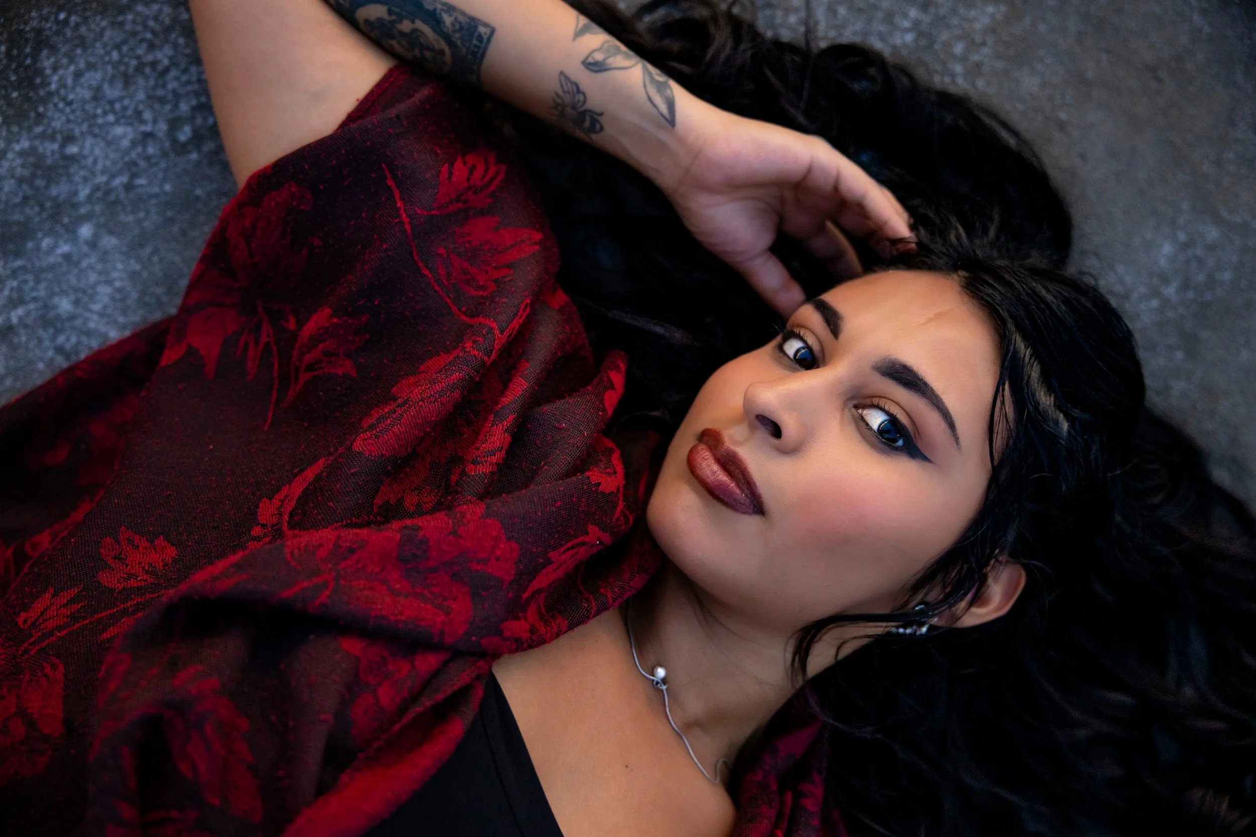 A woman with black hair and tattoos lying on a gray surface, wearing red and black floral clothing and looking at the camera.