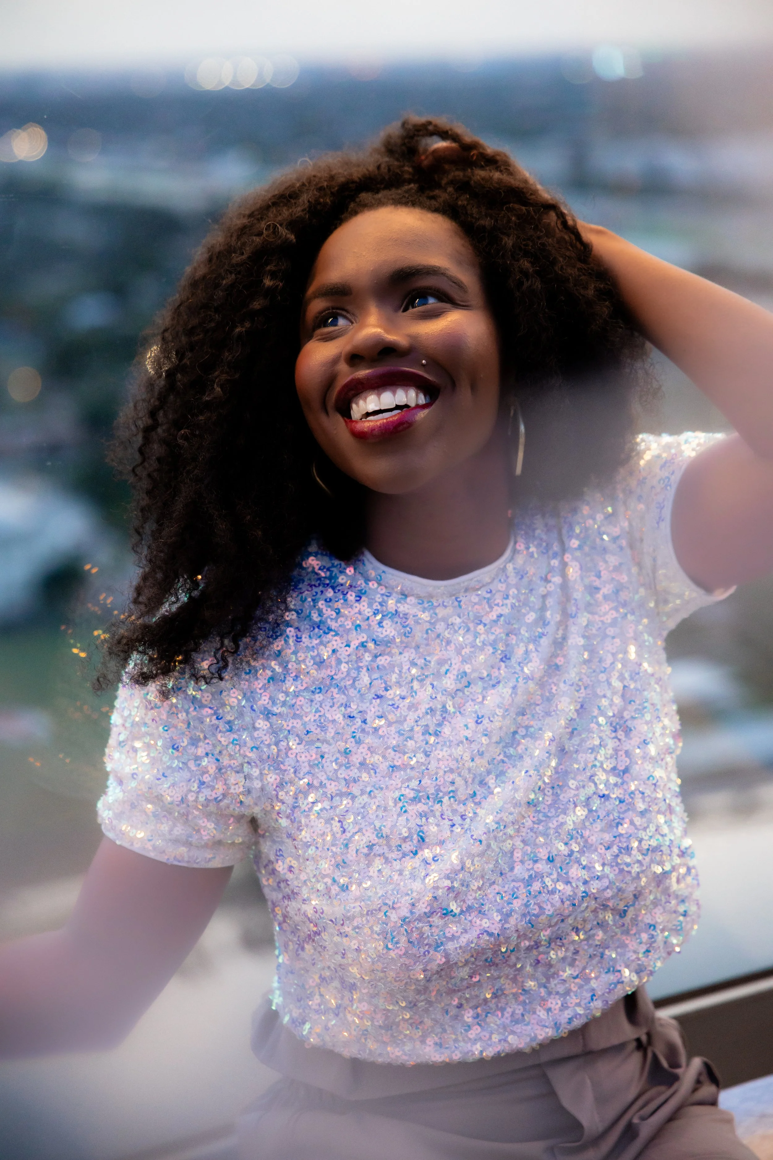 Smiling woman with curly hair, wearing a sparkly top, at the beach during sunset with bokeh lights in the background.