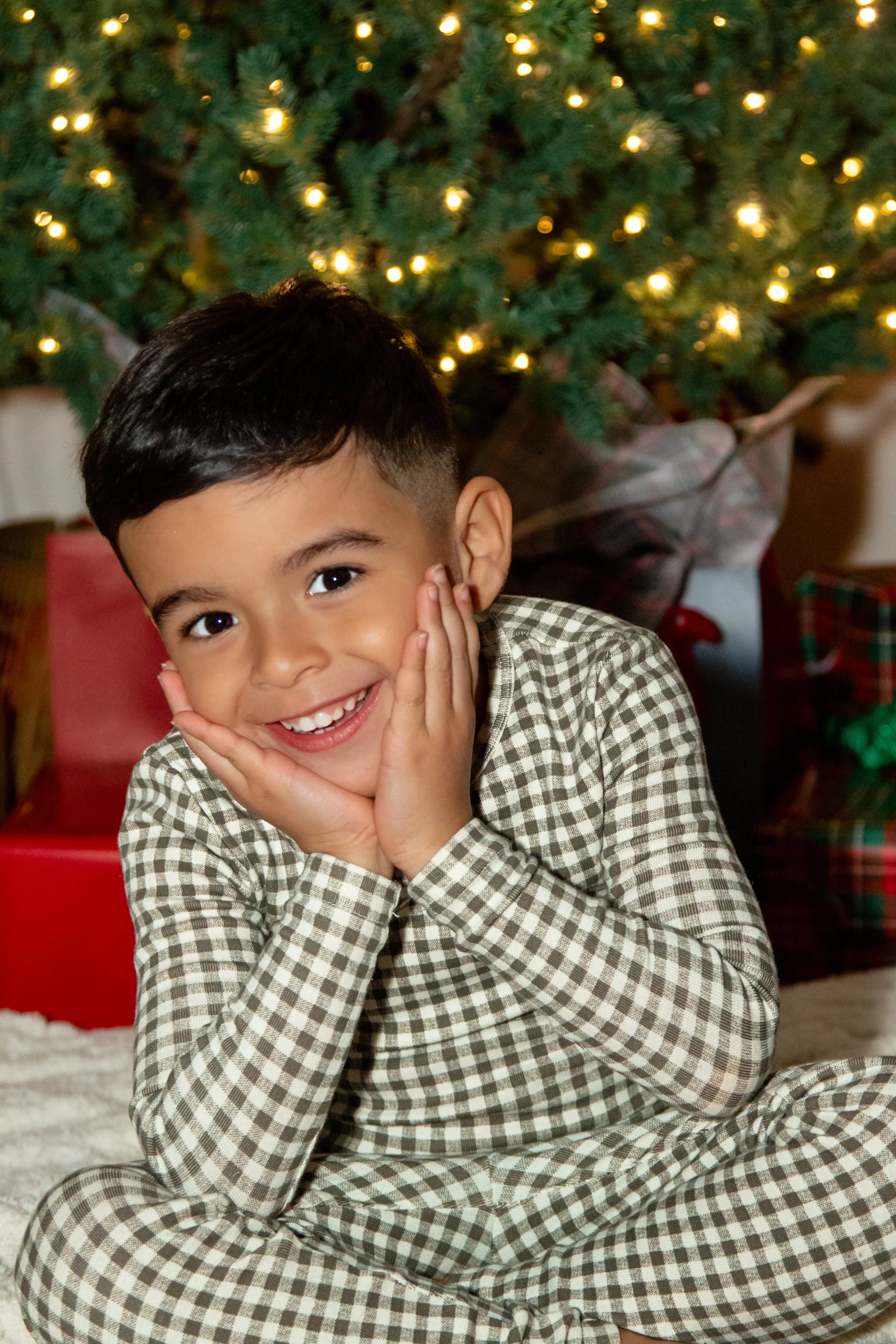 Young boy sitting in front of a decorated Christmas tree, smiling with his hands on his face.