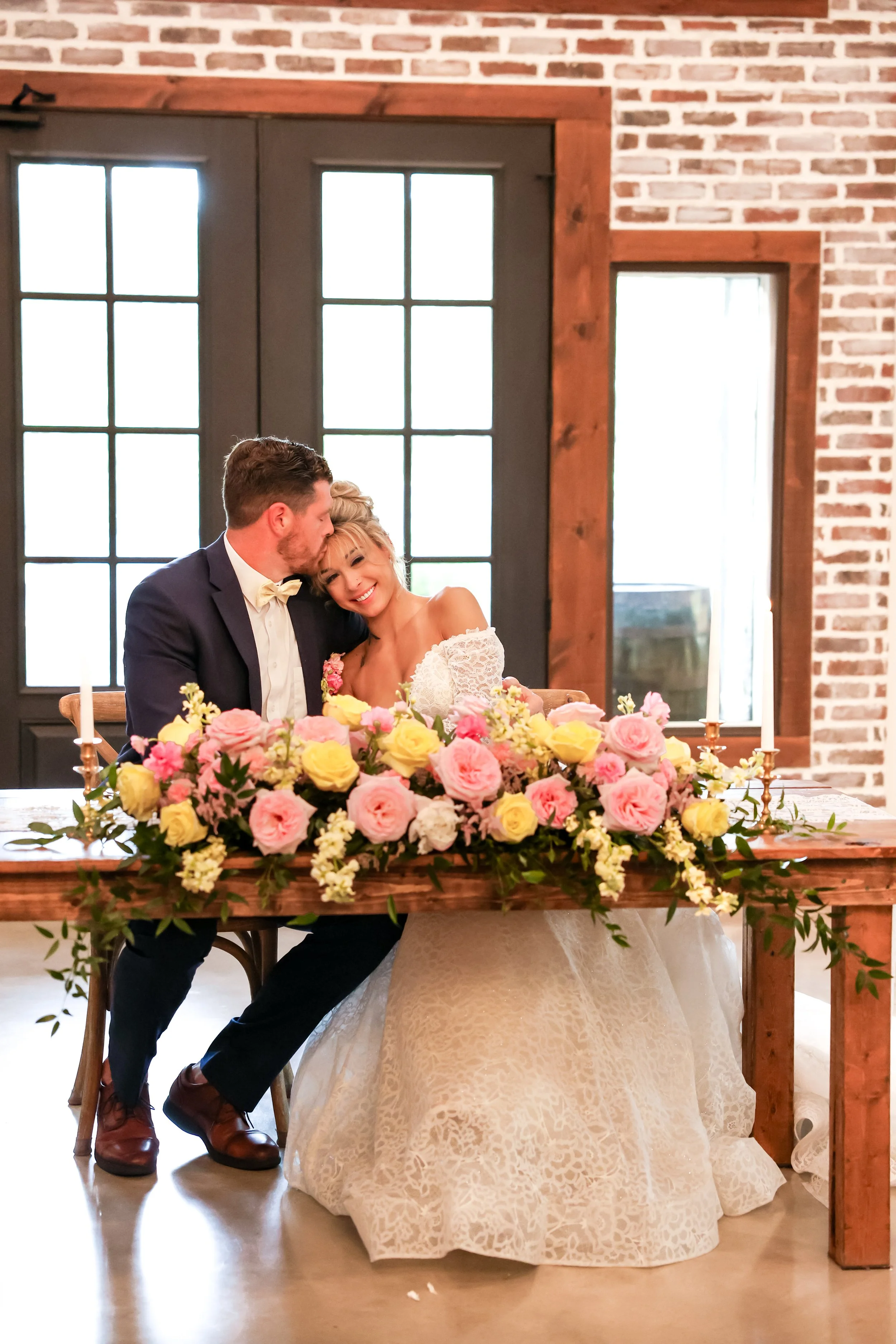 A bride and groom sit at a wedding reception table adorned with a large floral arrangement of pink and yellow roses and white flowers. The groom is whispering into the bride's ear, and she is smiling happily. The background features brick walls and a