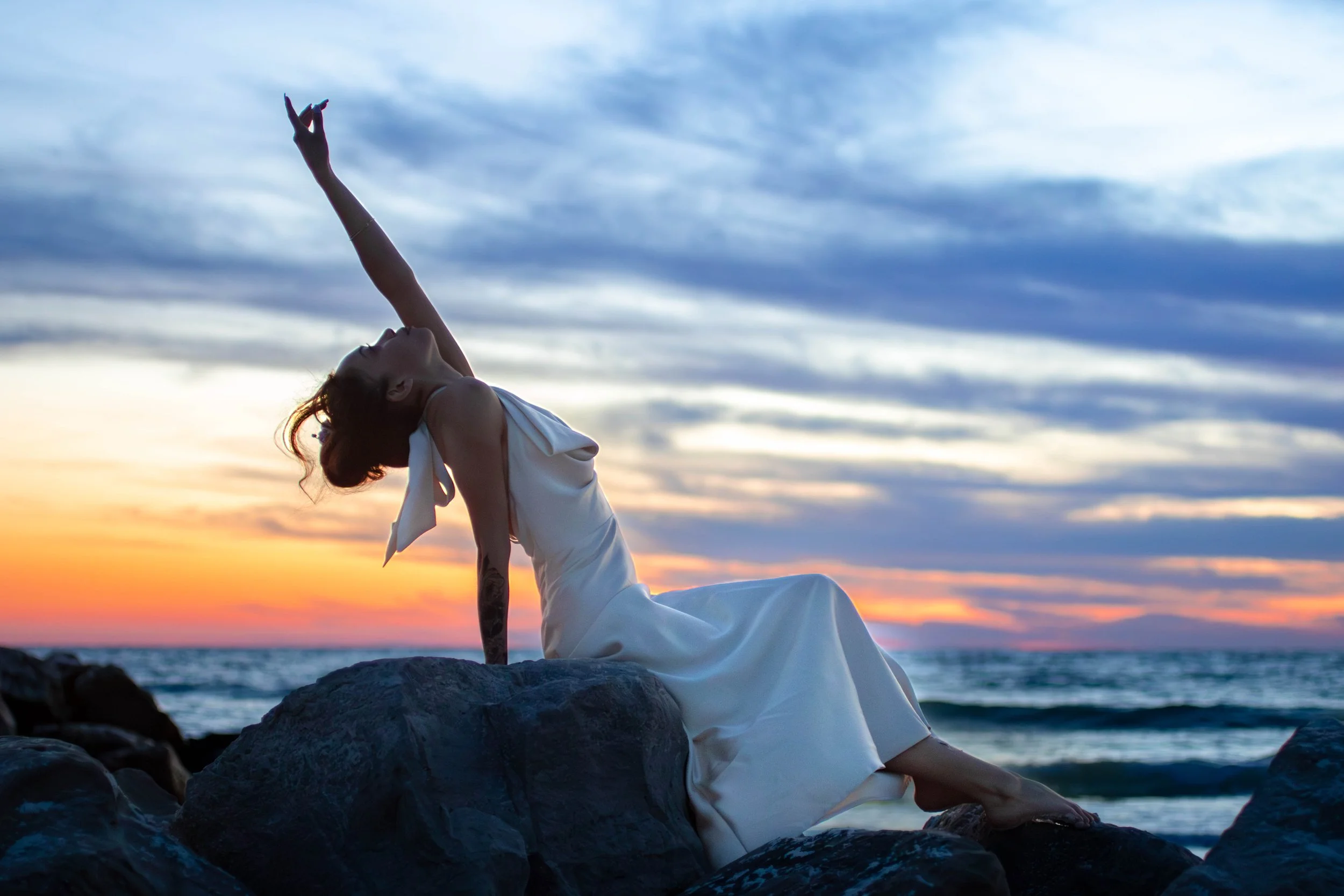 A woman practicing yoga on rocks by the ocean at sunset, in a white dress, with her head tilted back and one arm extended upward.