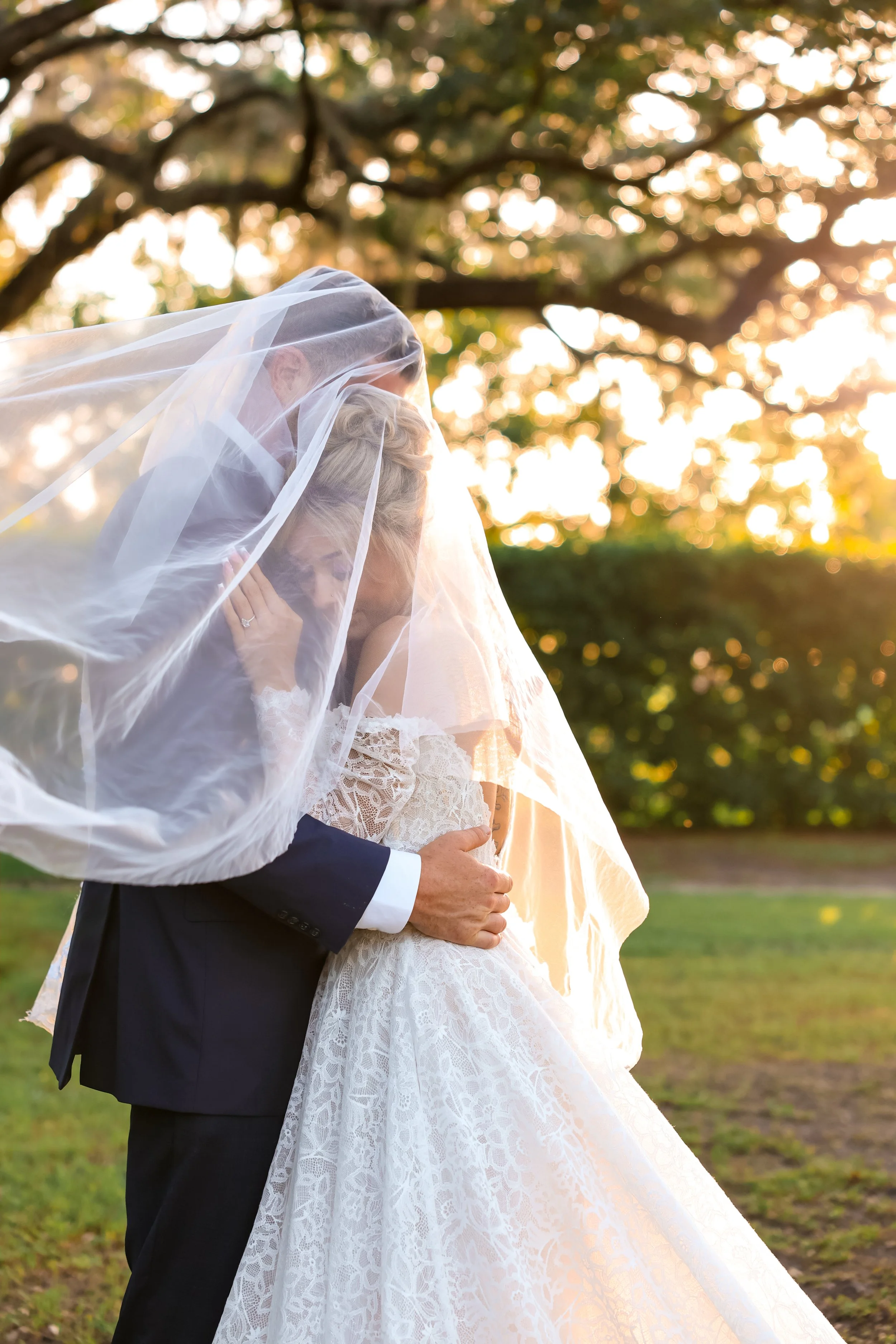 A bride and groom embracing outdoors during sunset, with the bride wearing a white lace wedding dress and veil, and the groom in a black tuxedo, in front of a tree with golden sunlight.