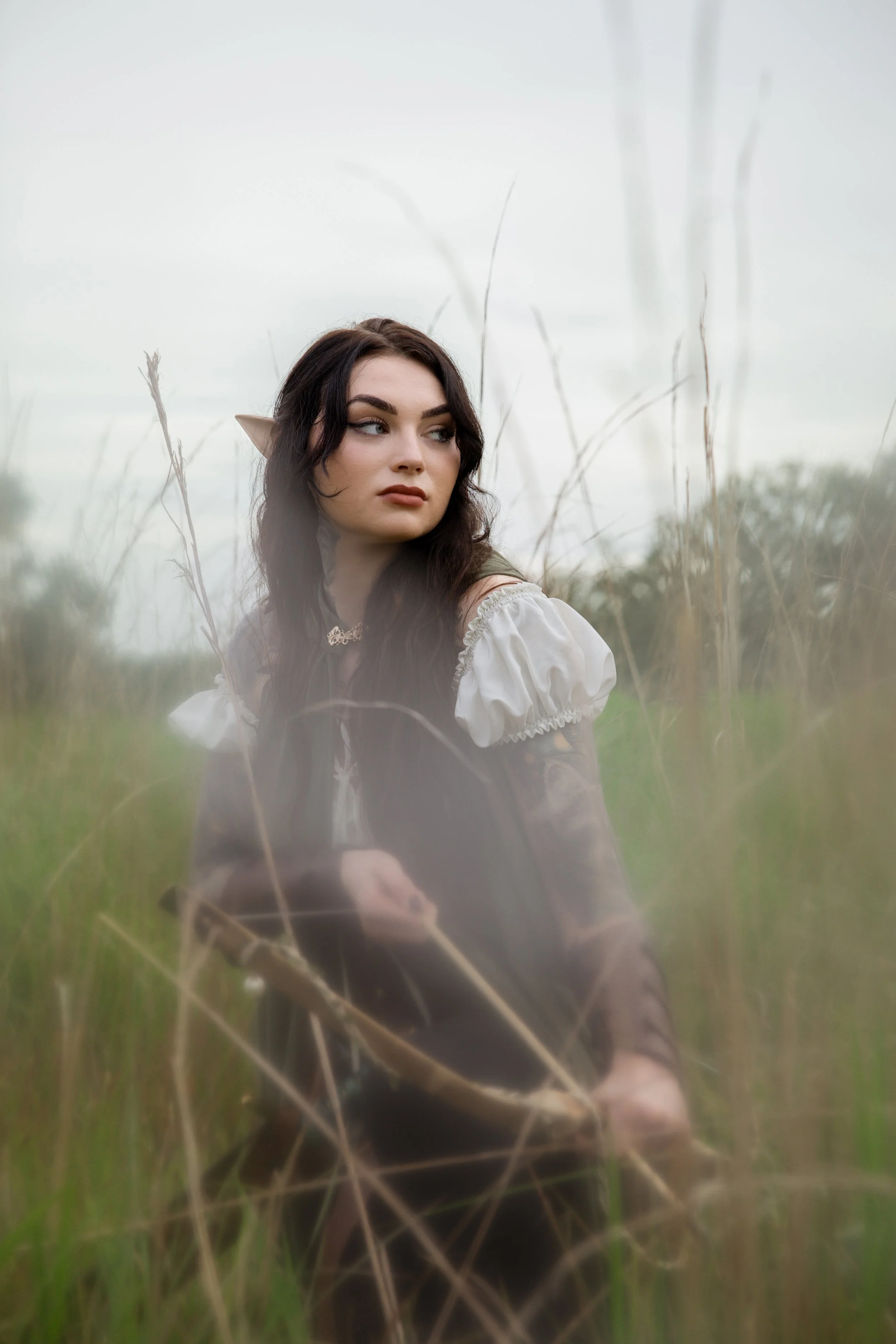 A Lord of the Rings ranger or archer inspired photo of a woman with elf ears and dark curly hair sitting in a field of tall grass, looking to the side with a neutral expression.