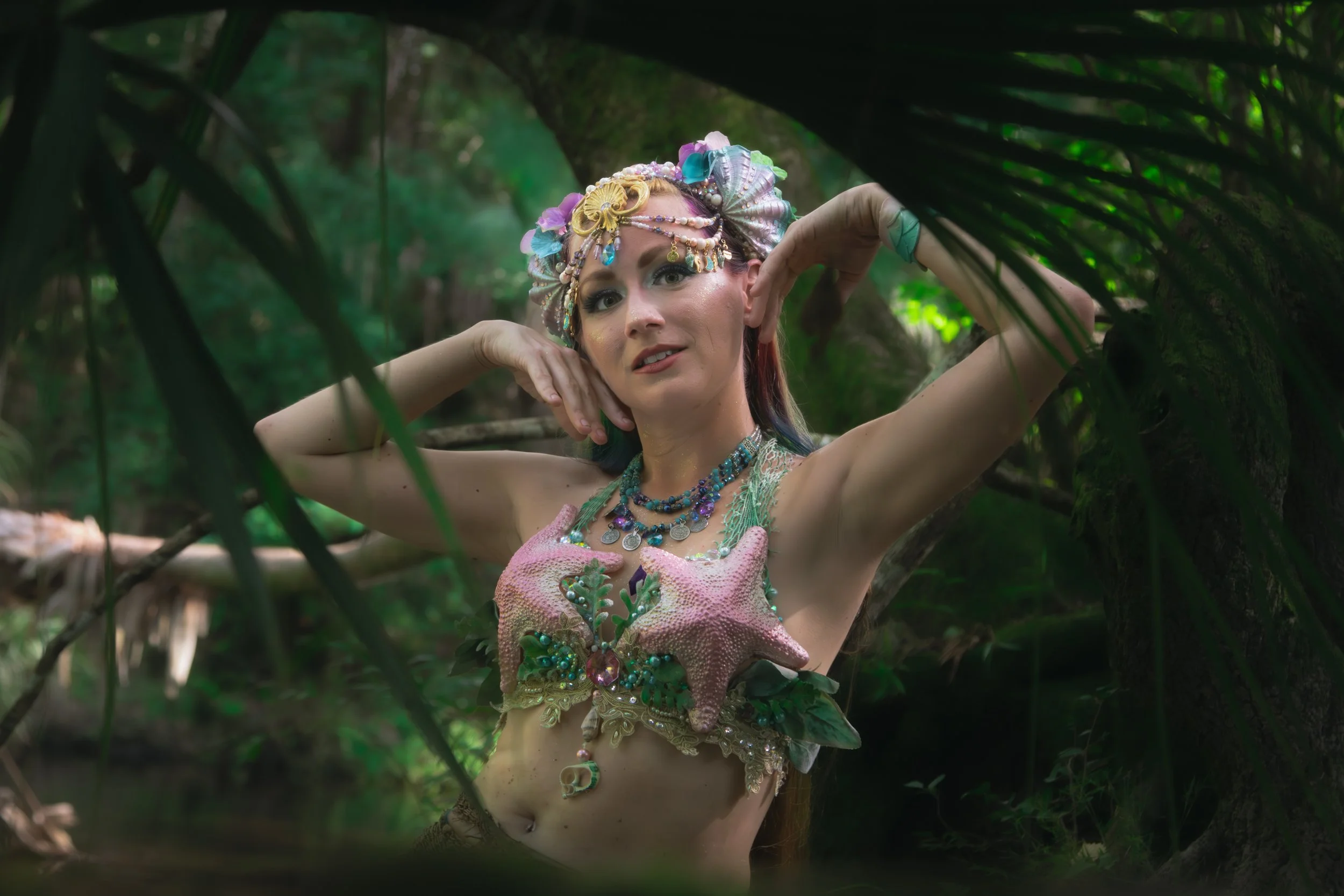 A woman dressed as a sea mermaid, wearing elaborate jewelry and a colorful shell top, poses in a lush, green jungle setting framed by dense foliage.