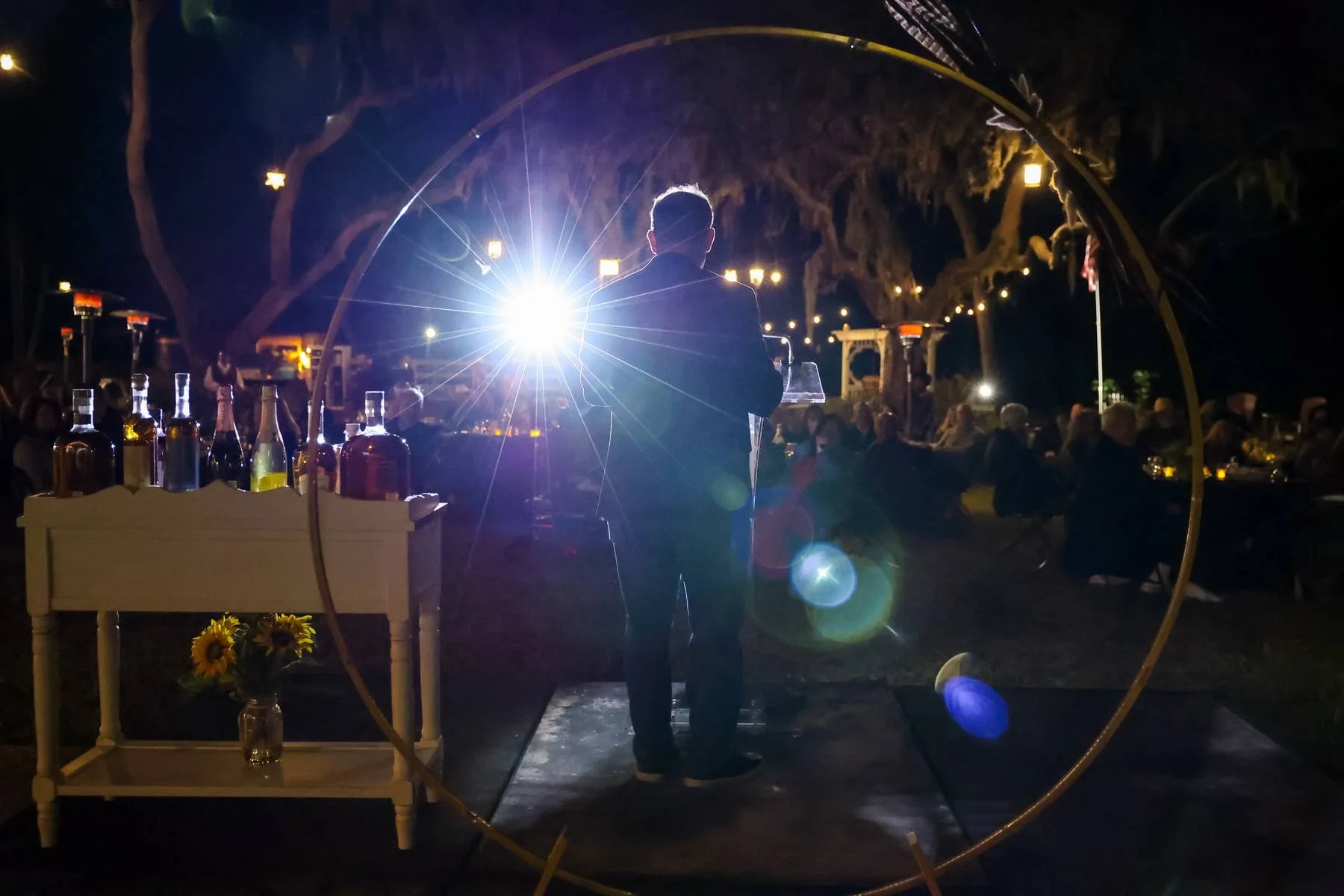 A person stands behind a DJ booth at an outdoor nighttime event, with a bright light behind them. The scene is decorated with hanging lights, and tables with people sitting, eating, and drinking. There are bottles on a nearby cart and a vase with sun