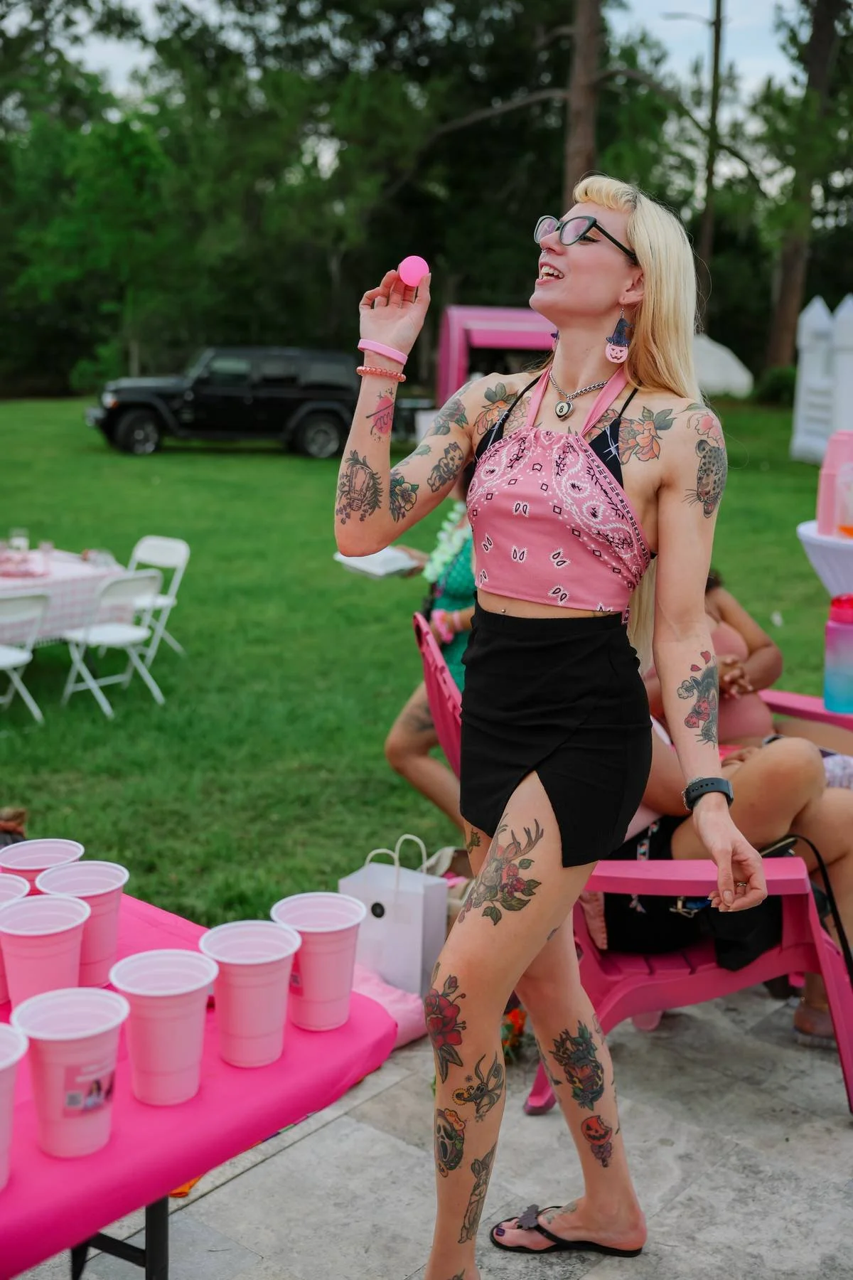 A woman with blonde hair and tattoos, wearing glasses, a pink bandana top, black skirt, and flip-flops, holding a pink cup and a ping-pong ball at an outdoor gathering with pink decorations and plastic cups on a table.