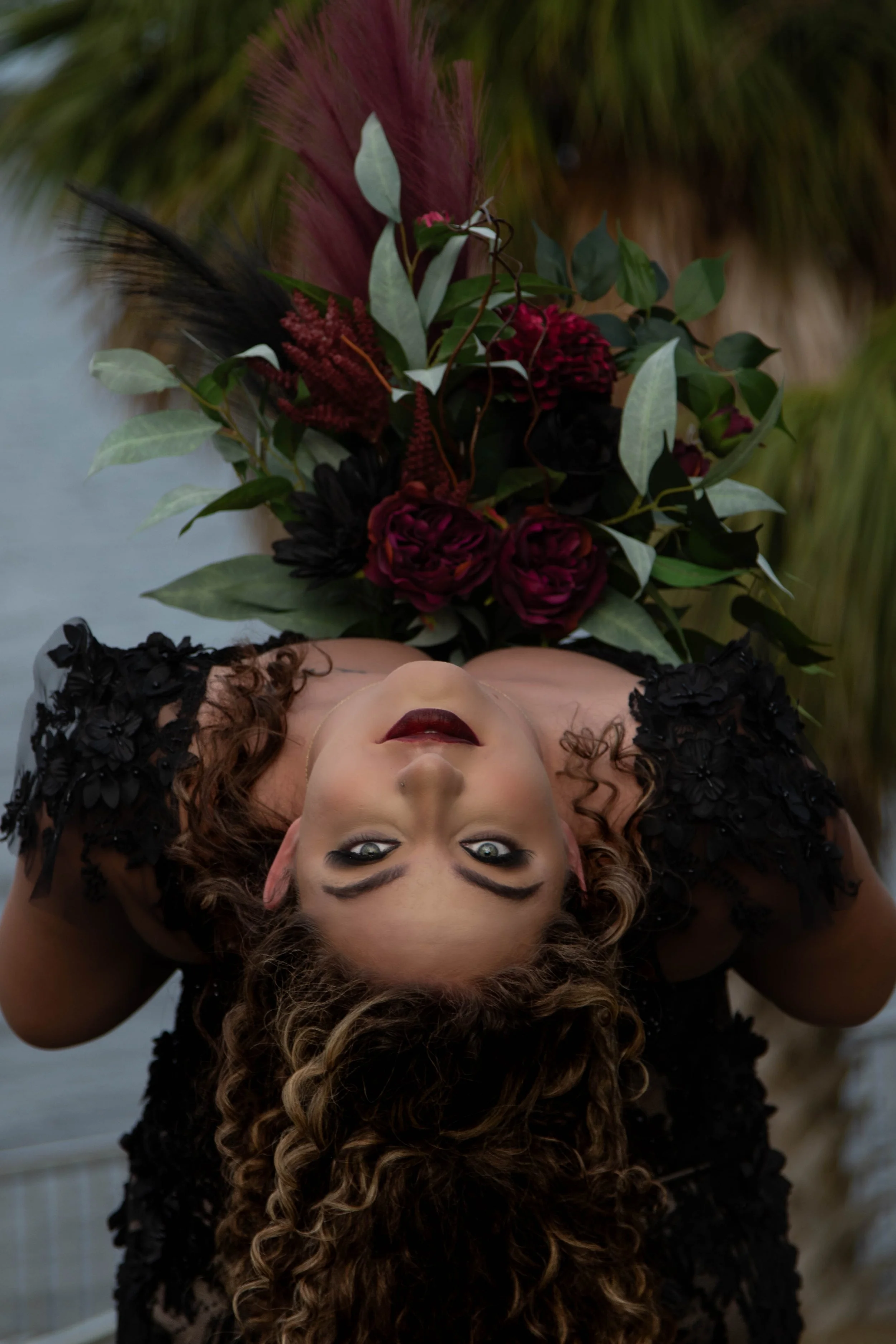 A woman with curly brown hair, dark eye makeup, and dark red lipstick lying upside down with her head on the ground. She is wearing a black dress with floral embellishments and is holding a bouquet of dark red and pink flowers, including roses and ot