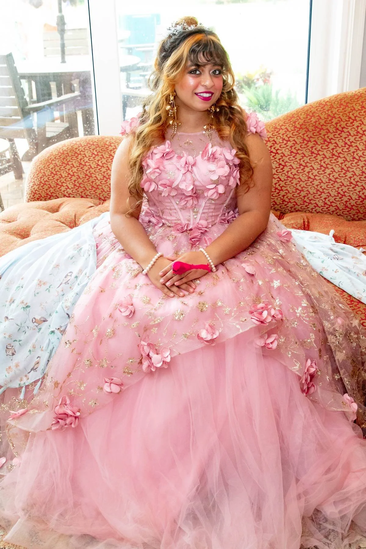 Young woman wearing a pink floral-embellished ball gown, sitting on a red patterned sofa in front of a glass door with an outdoor patio visible behind her.