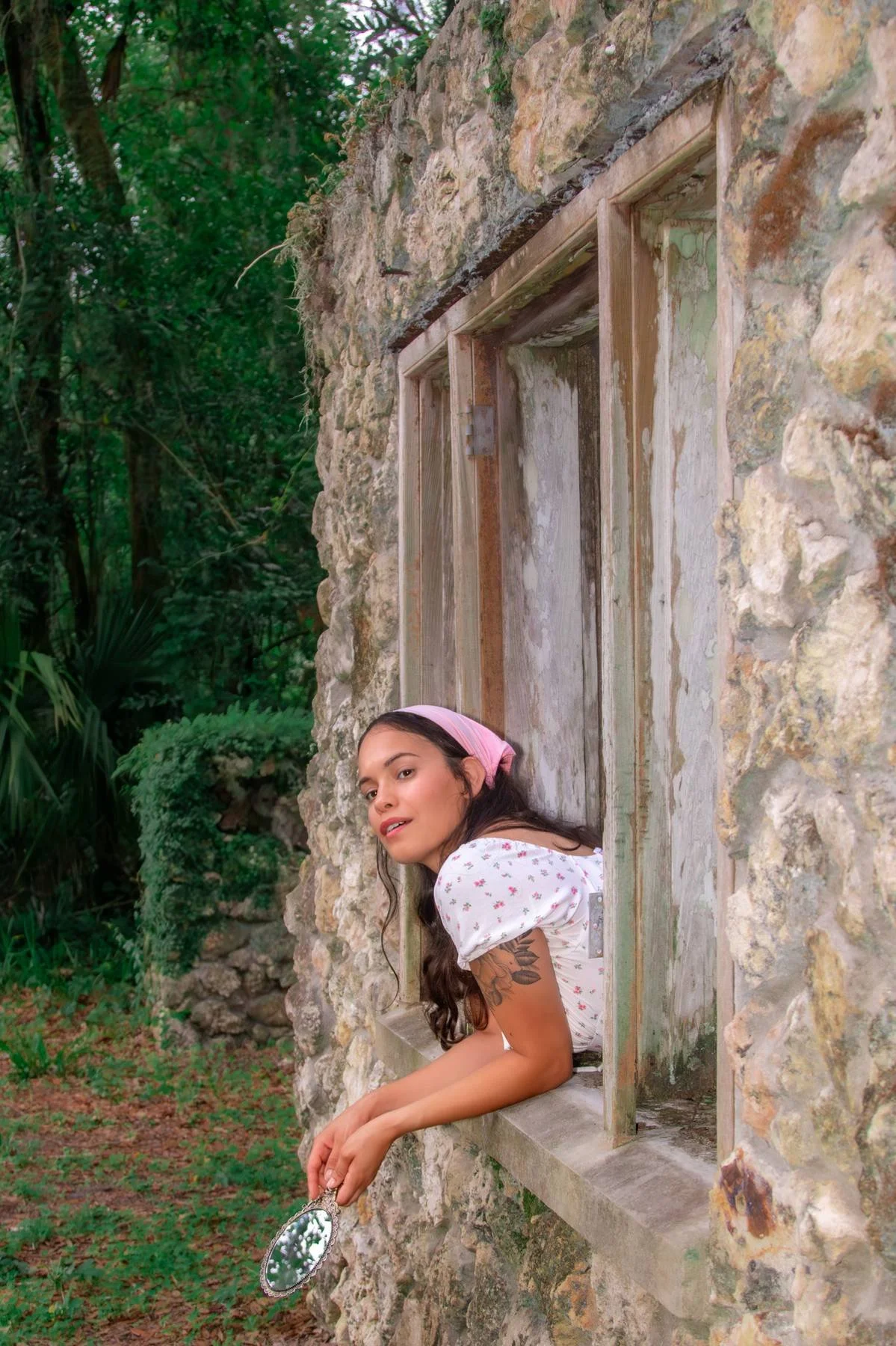 Young woman with long dark hair, wearing a pink headscarf and a white floral dress, leaning out of a stone window of an old building surrounded by greenery, holding a small mirror.